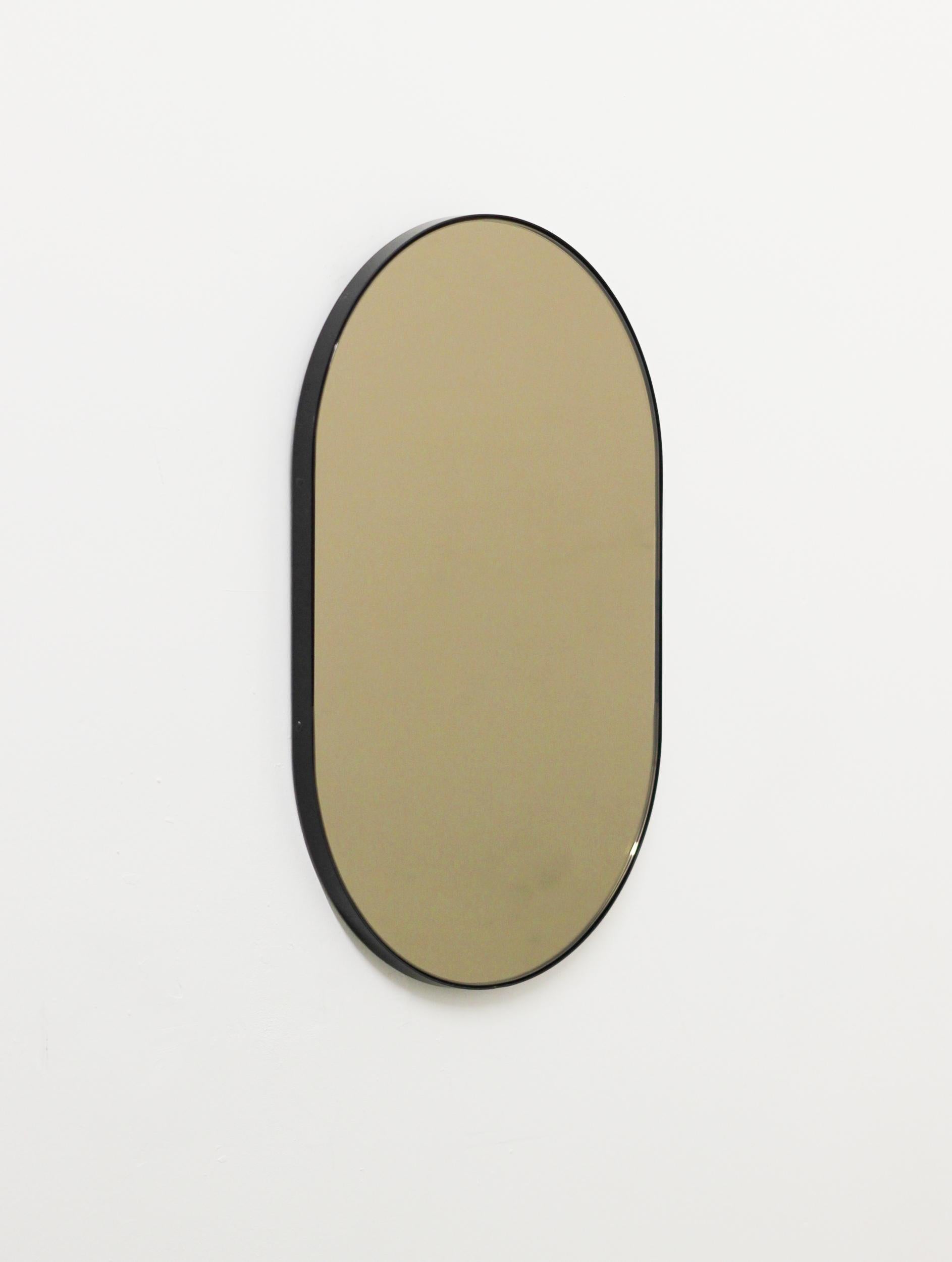 Moderner, handgefertigter, kapselförmiger, bronzefarben getönter Spiegel mit einem eleganten, schwarz pulverbeschichteten Aluminiumrahmen. Entworfen und handgefertigt in London, UK.

Je nach Größe des Spiegels mit einem Messinghaken oder einer