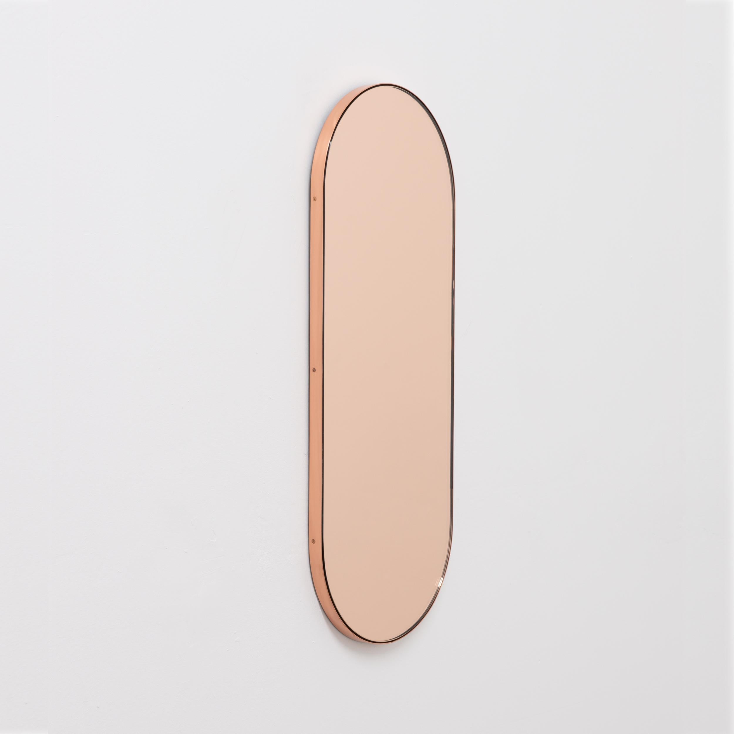 Contemporary capsule shaped rose gold or peach mirror with an elegant copper frame. Designed and handcrafted in London, UK. The detailing and finish, including visible copper-plated screws, emphasise the craft and quality feel of the mirror, a true