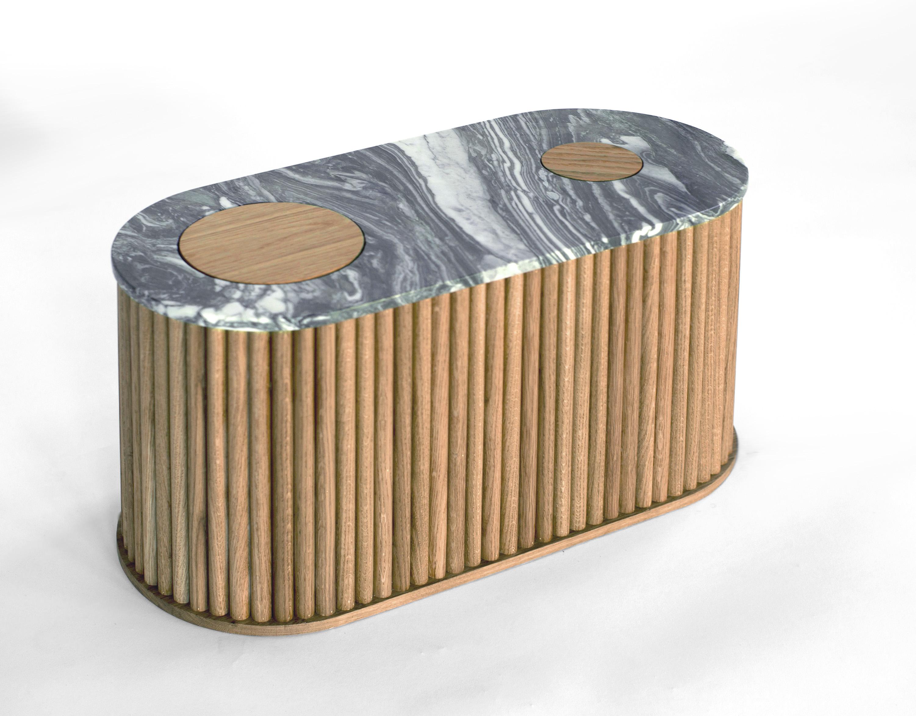 Cette table de chevet est disponible individuellement ou en set de deux. Son plateau est arrondi de chaque côté avec des découpes circulaires insérées révélant la matérialité de sa base en bois. La base est enveloppée d'un tambour en chêne blanc