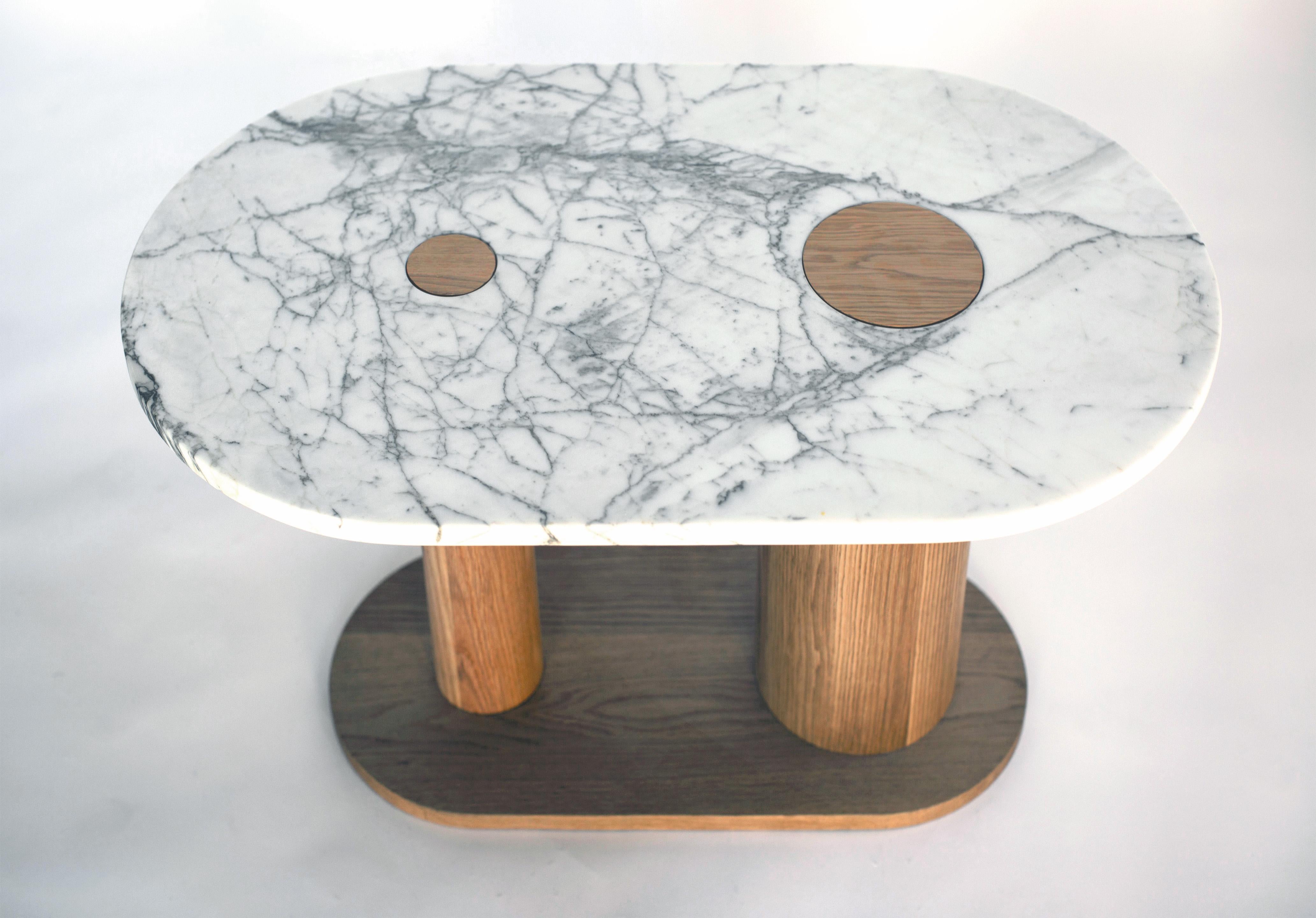 This coffee table comes in a striking two-toned marble, cream white with veining in dark gray with green undertones. Two cutouts in the marble top reveal the materiality of the asymmetrical legs below. The legs, made in solid white oak, are