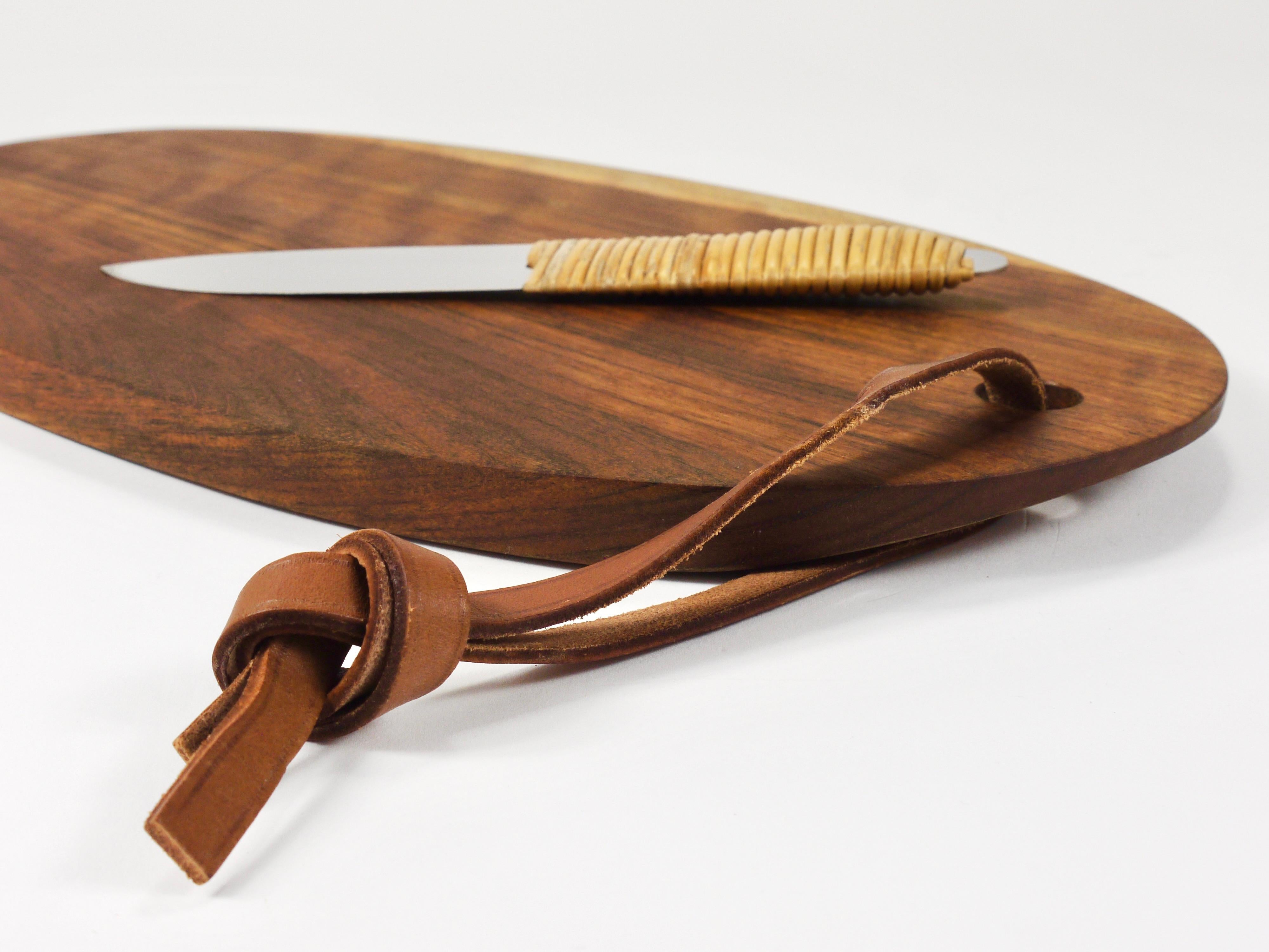 A beautiful oval chopping board or serving tray with a knife, designed and handcrafted in the 1950s by Werkstätte Carl Auböck. The cutting board is made of beautifully grained walnut and features a leather strap, showcasing Auböck's renowned