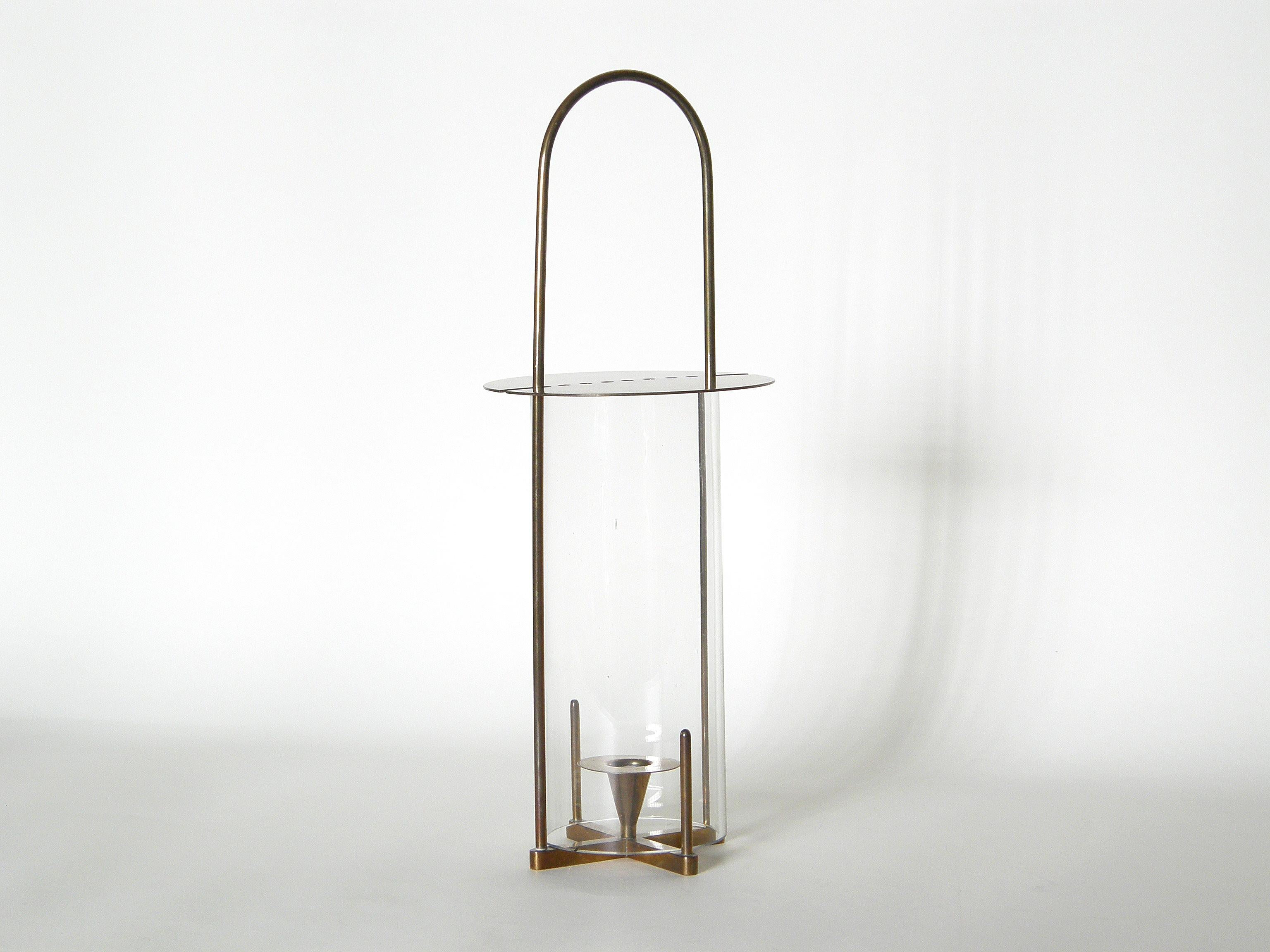 This Carl Auböck candle lantern has a hurricane type, cylindrical glass shade. The top of the brass frame forms a handle, and the brass cap has a line of small holes to allow air flow for the candle.

The height given is to the top of the