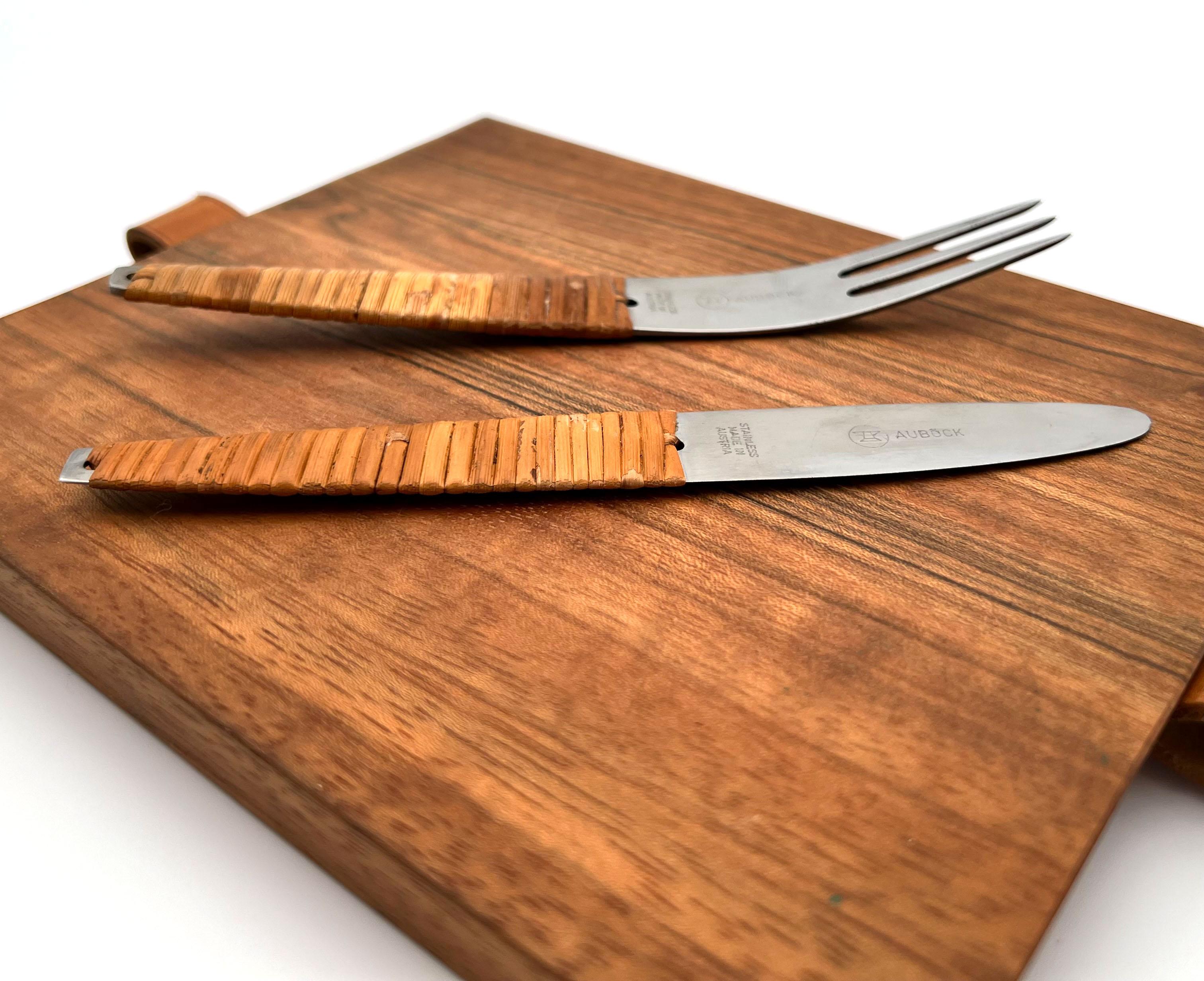 Carl Aubock Cutting Board & Knife and Fork Set. 

Original detailing and finishes, walnut cutting board, stainless steel flatware, rattan wrapped handles, leather strap.

Stamped signature to blade: “Auböck