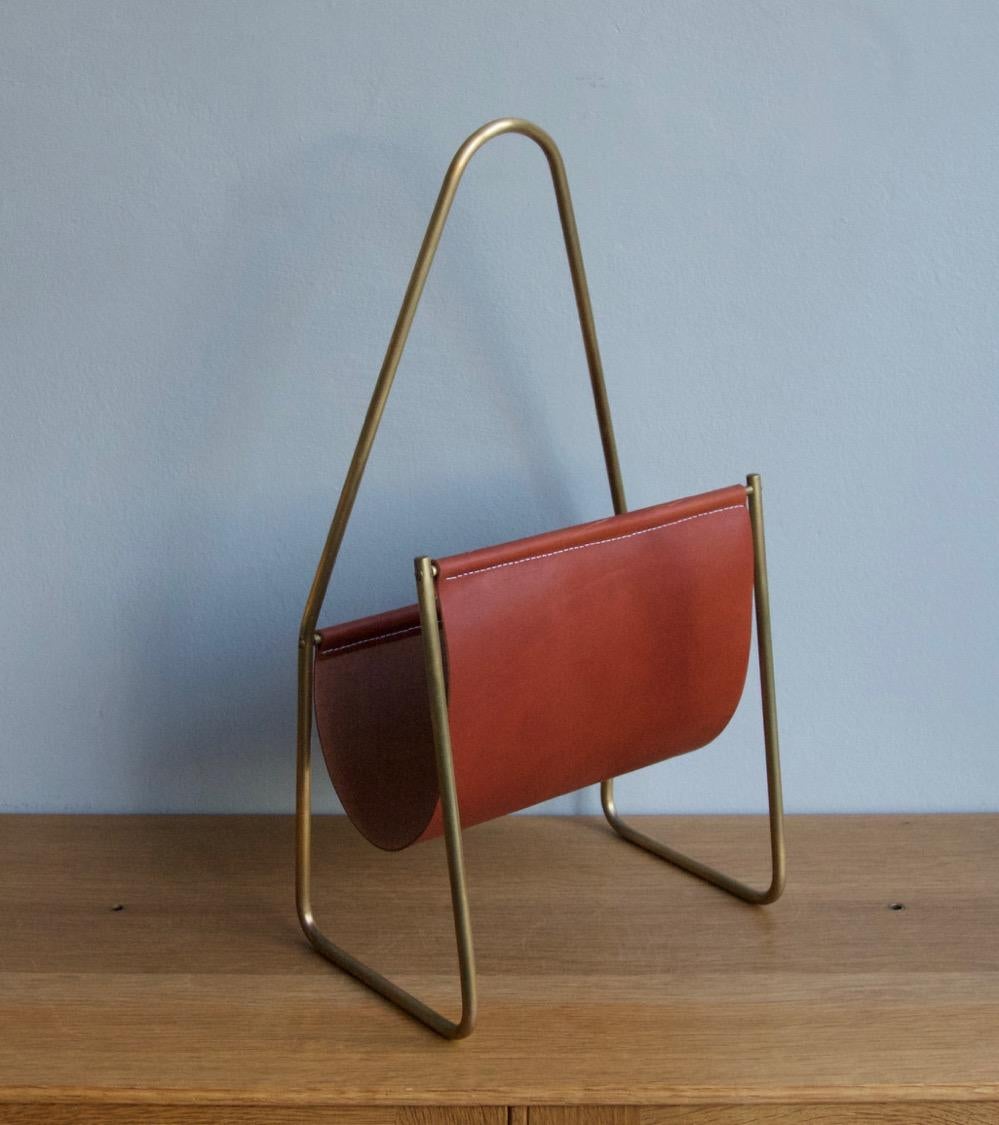 Magazine holder designed and made in the Auböck Werkstatte, Vienna, circa 1960. 
Numbered as design 3808 of the Viennese workshop, it features a sinuous frame in brushed brass covered by a uniform patina and a leather sling with white