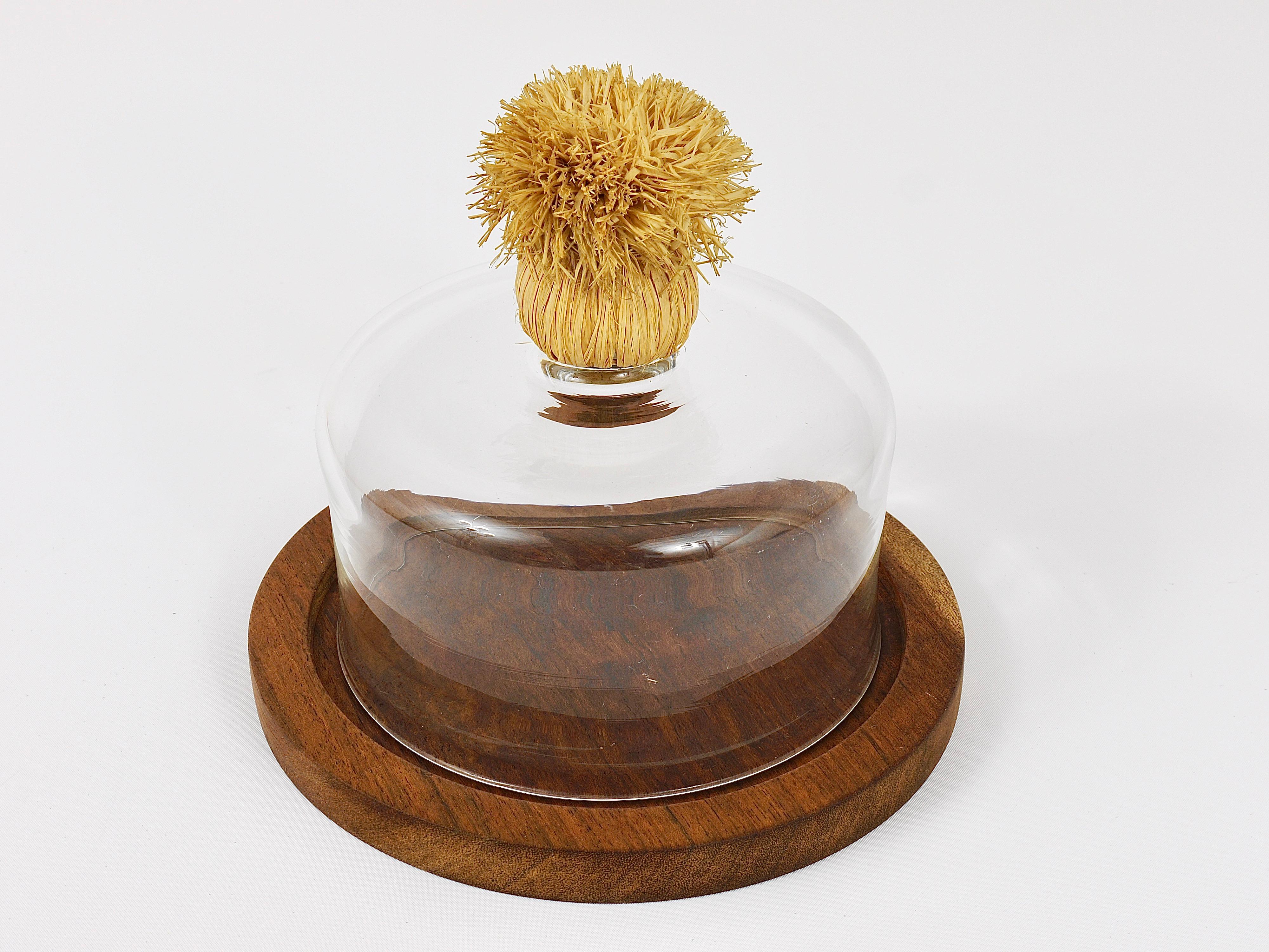 A beautiful midcentury walnut cheese board with a glass dome and a raffia handle from the 1950s. Designed and executed by Carl Auböck. The total diameter is 8 inches, the glass dome has a diameter of 6 inches and a height of 3 1/2 inches without the