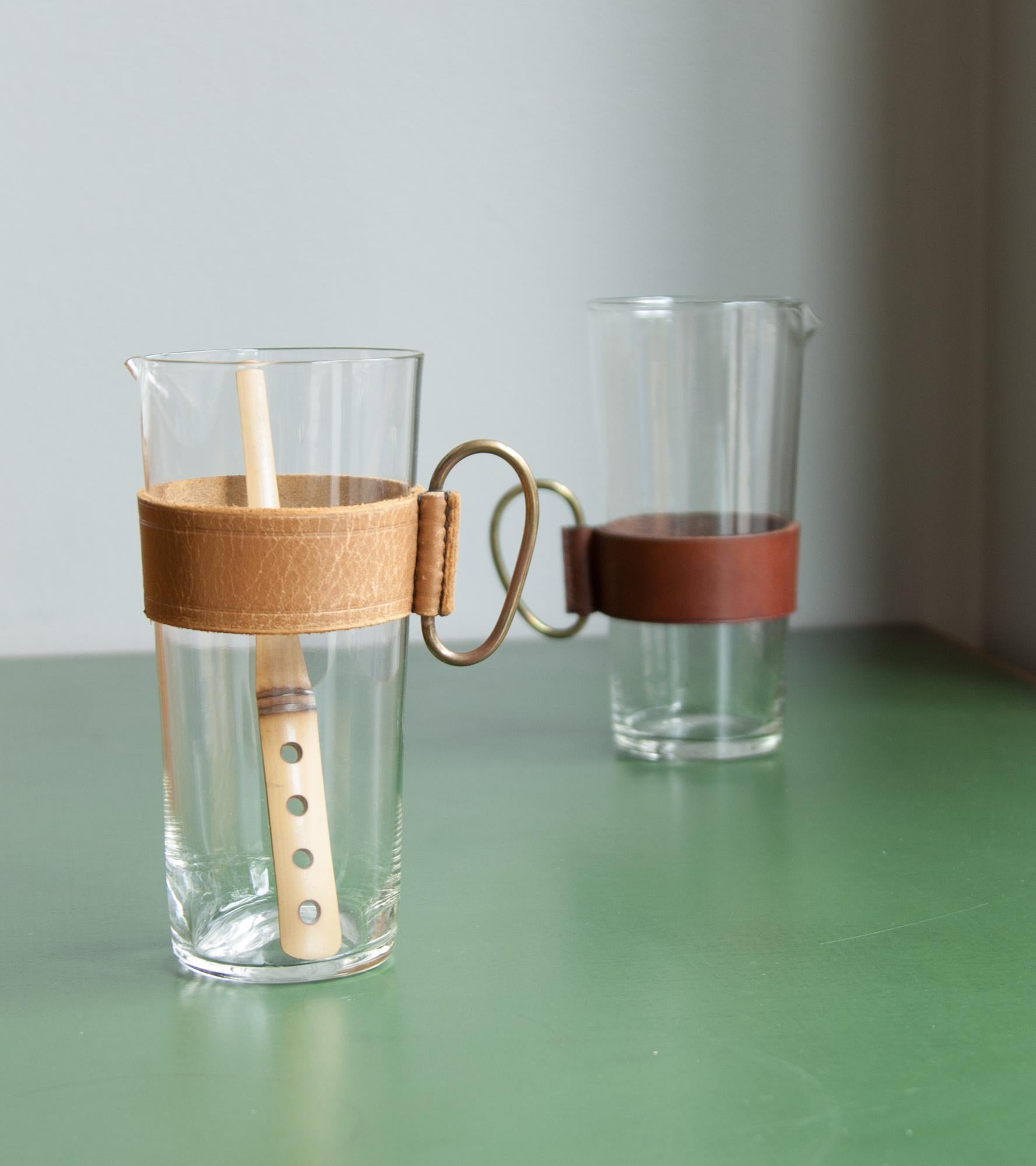 A glass pitcher with leather sleeve and brass handle by Carl Auböck II, Vienna, 1950s.
The leather sleeve settles in the bottom half of the glass and is of a deep dark brown colour. The brass handle has a uniform warm patina naturally built