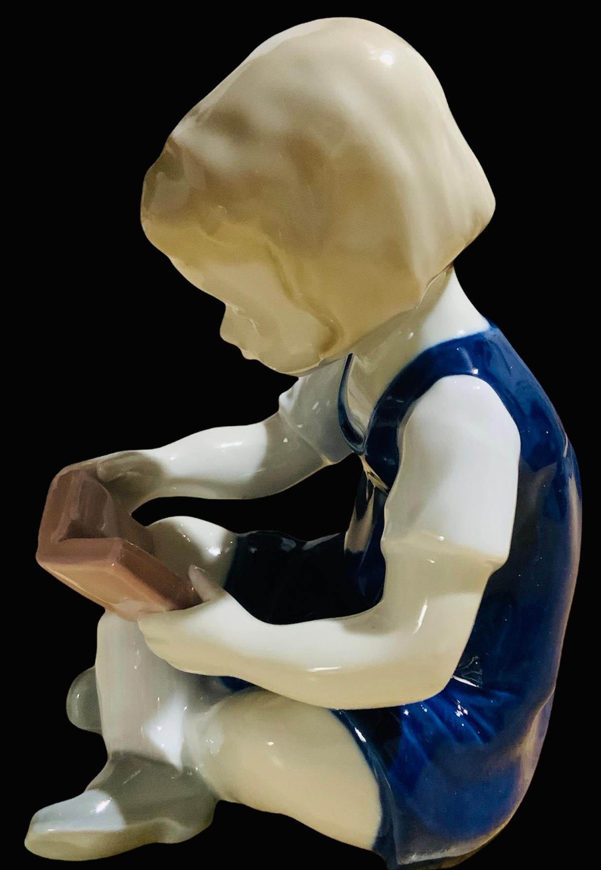 This is a Carl Scheidig German porcelain figurine of a girl reading a book. It depicts a girl dressed with a blue jumper, white shirt and grey shoes seated with her legs crossed, holding a book that she is reading. Below the base, it is found the