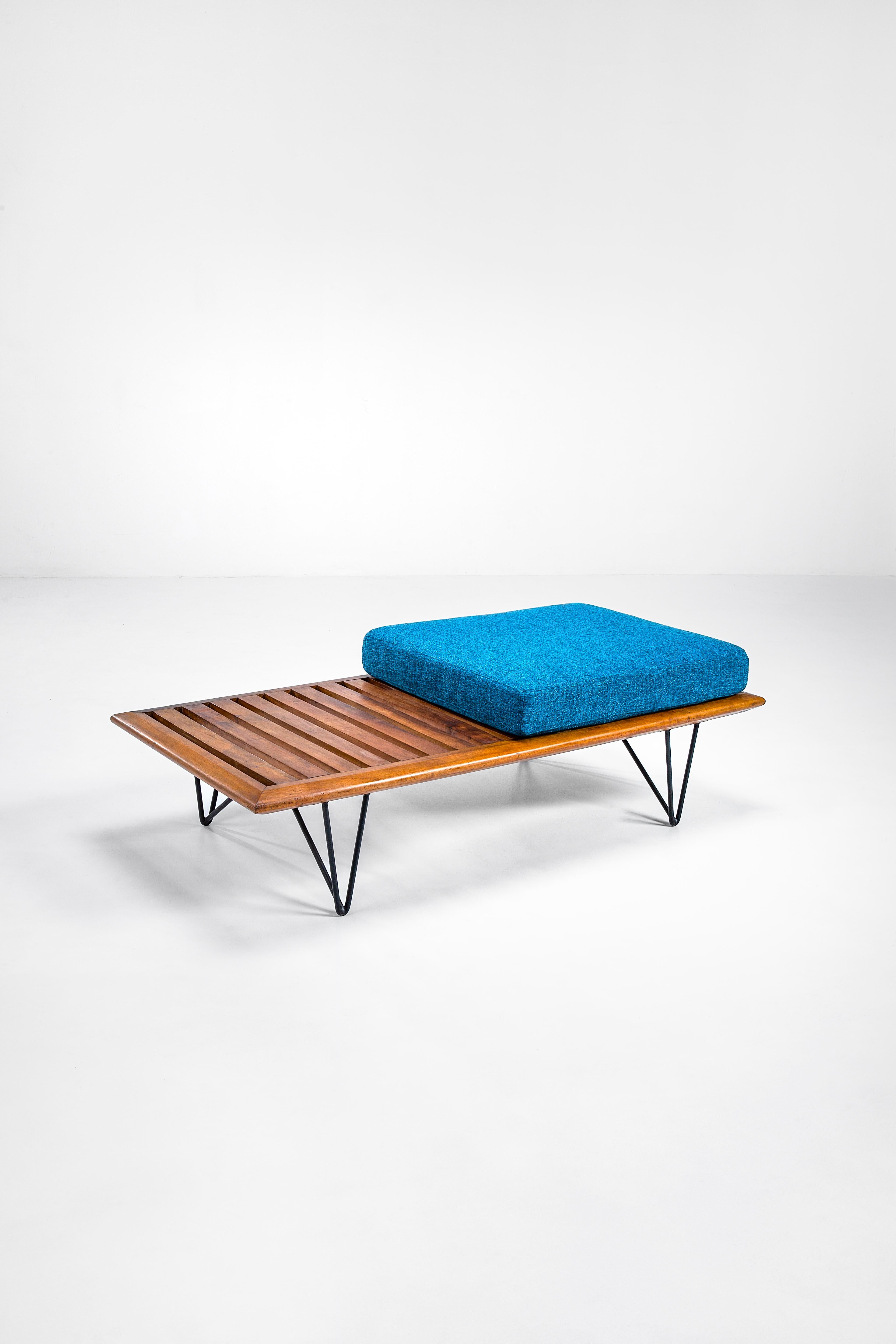 This delicate bench with a suspended seat on leather or fabric strips and a thick fabric cushion is accompanied by a section that functions as a tabletop. The overall structure is wood, with tubular metal supports. The forms thus trace the