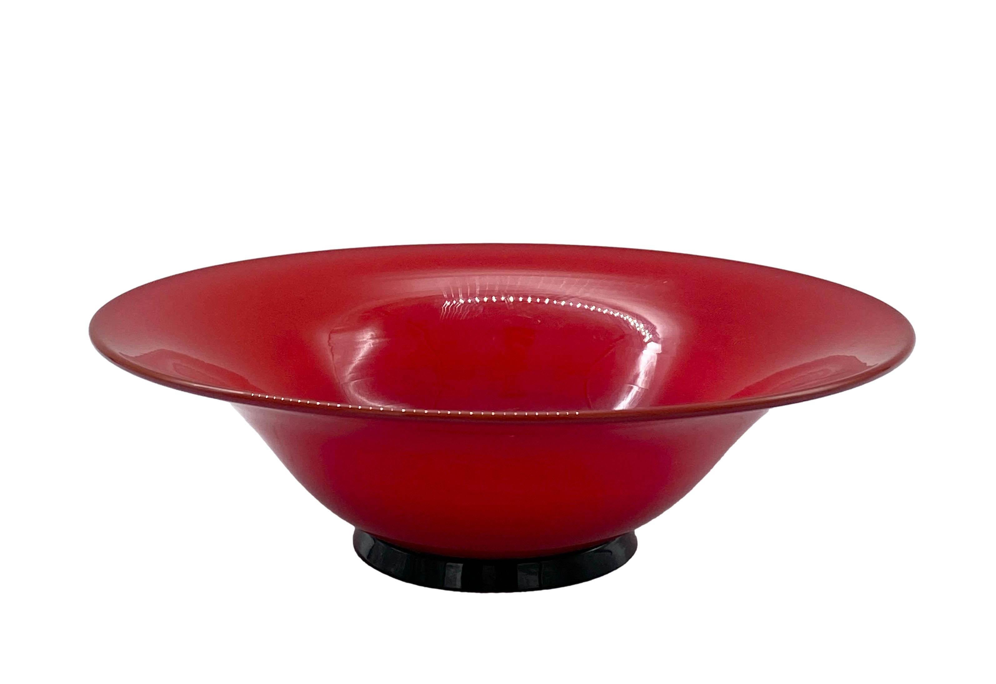 Bowl in red and milky coated Murano glass, produced by the famous artistic glassworks Venini, based on the collection of the 1930s designed by Carlo Scarpa. Inside, the red glass takes on a warm, amber hue. It rests on a small circular black glass