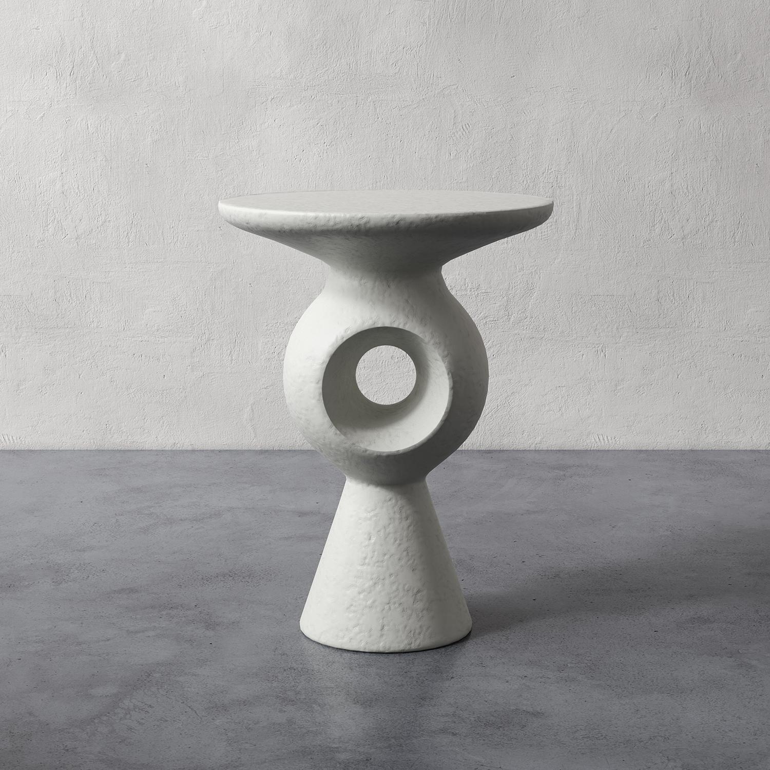 Esta mesa auxiliar con pedestal de escayola presenta una silueta tallada única, con un recorte en el centro que añade un toque artístico a cualquier espacio. Inspirada en las obras de los escultores Jean Arp y Barbara Hepworth, la forma orgánica de