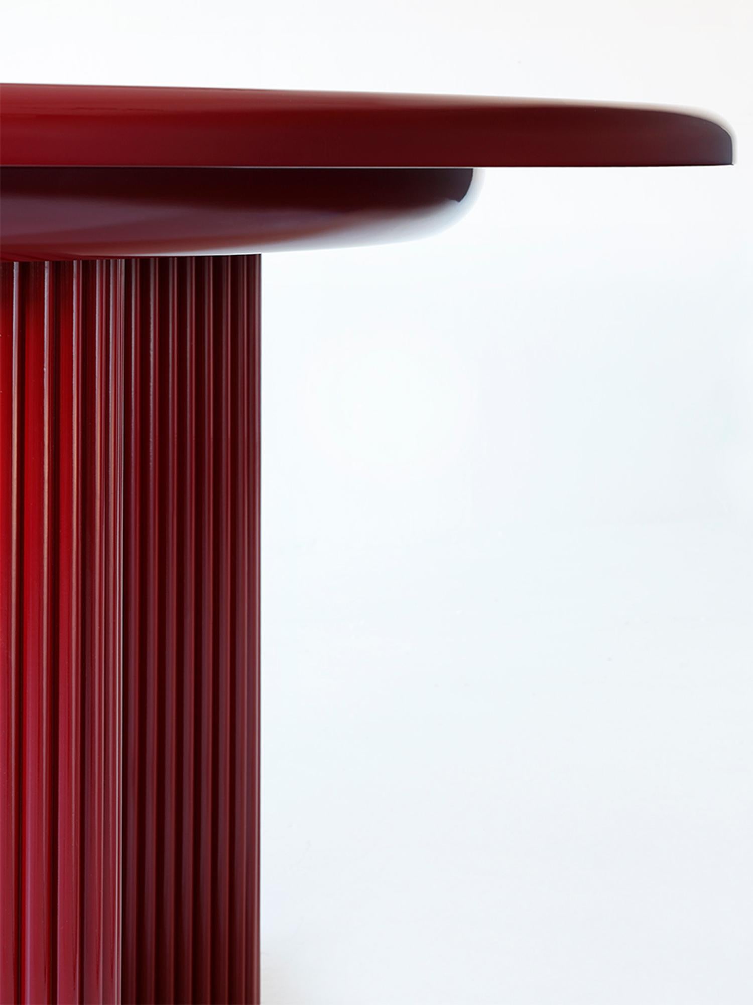 Un tavolo da pranzo scultoreo definito dalla sua forma audace e dai suoi dettagli raffinati. Le gambe in legno a coste, rifinite in lacca rossa, poggiano su uno zoccolo in ottone lucido. Il piano del tavolo laccato lucido è liscio al tatto, con