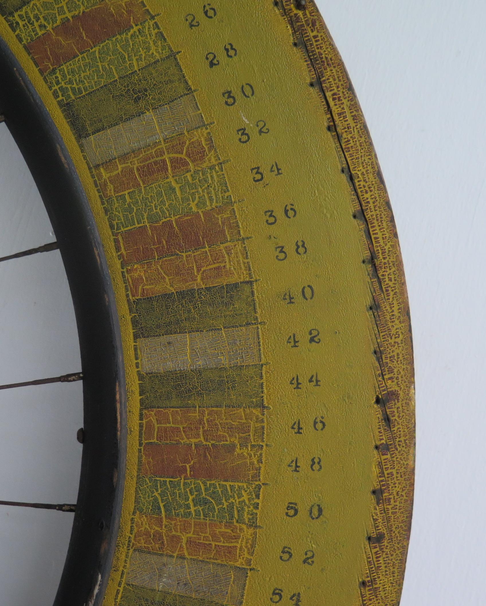 Carnival gaming wheel made from a bicycle wheel joined to a painted wood ring. The wood is painted in bands of softened colors and numbers with an aged cracked surface. The back of the wheel has a circle of nails that would spin and be slowed by a