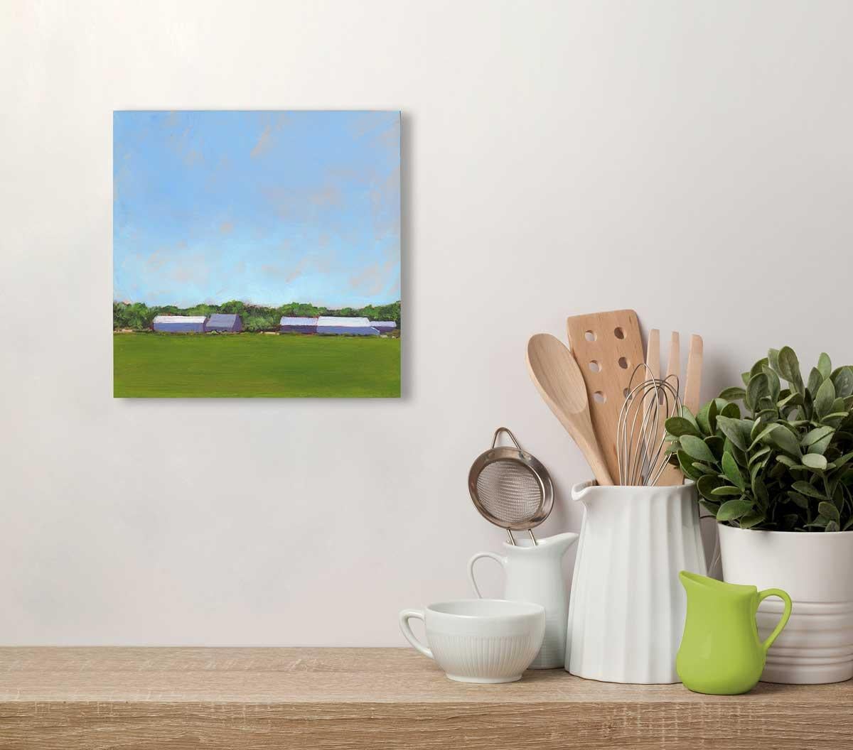 This small contemporary landscape painting by Carol Young features a scene of farmland, with green grass and foliage and cool violet barn structures along the horizon, beneath a painterly bright blue sky. The painting is made on board, and has clean