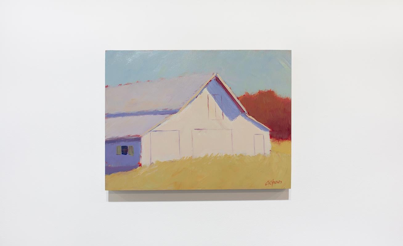 This small contemporary landscape painting by Carol Young features a warm neutral palette and captures a rural scene. A light-toned barn sits in a golden yellow field in front of deep red foliage, beneath a muted blue sky. The structure casts a