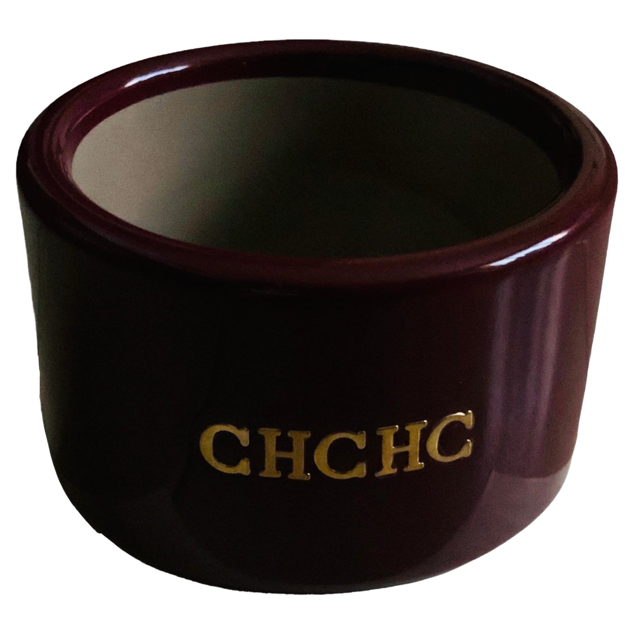 This is a Carolina Herrera cranberry color wide bangle. It is adorned in the center with gilt metal upper case initials- CHCHC from the designer name. 