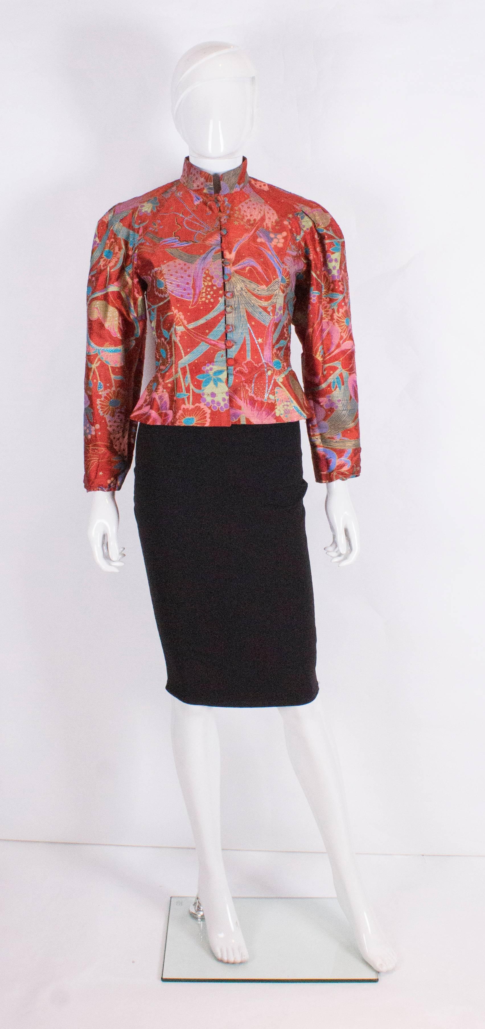 A pretty silk jacket by Caroline Charles. The jacket has a red background with a blue, gold and pink print. The jacket has a stand up collar, button front, gathered sleeves, and is flared at the waist.