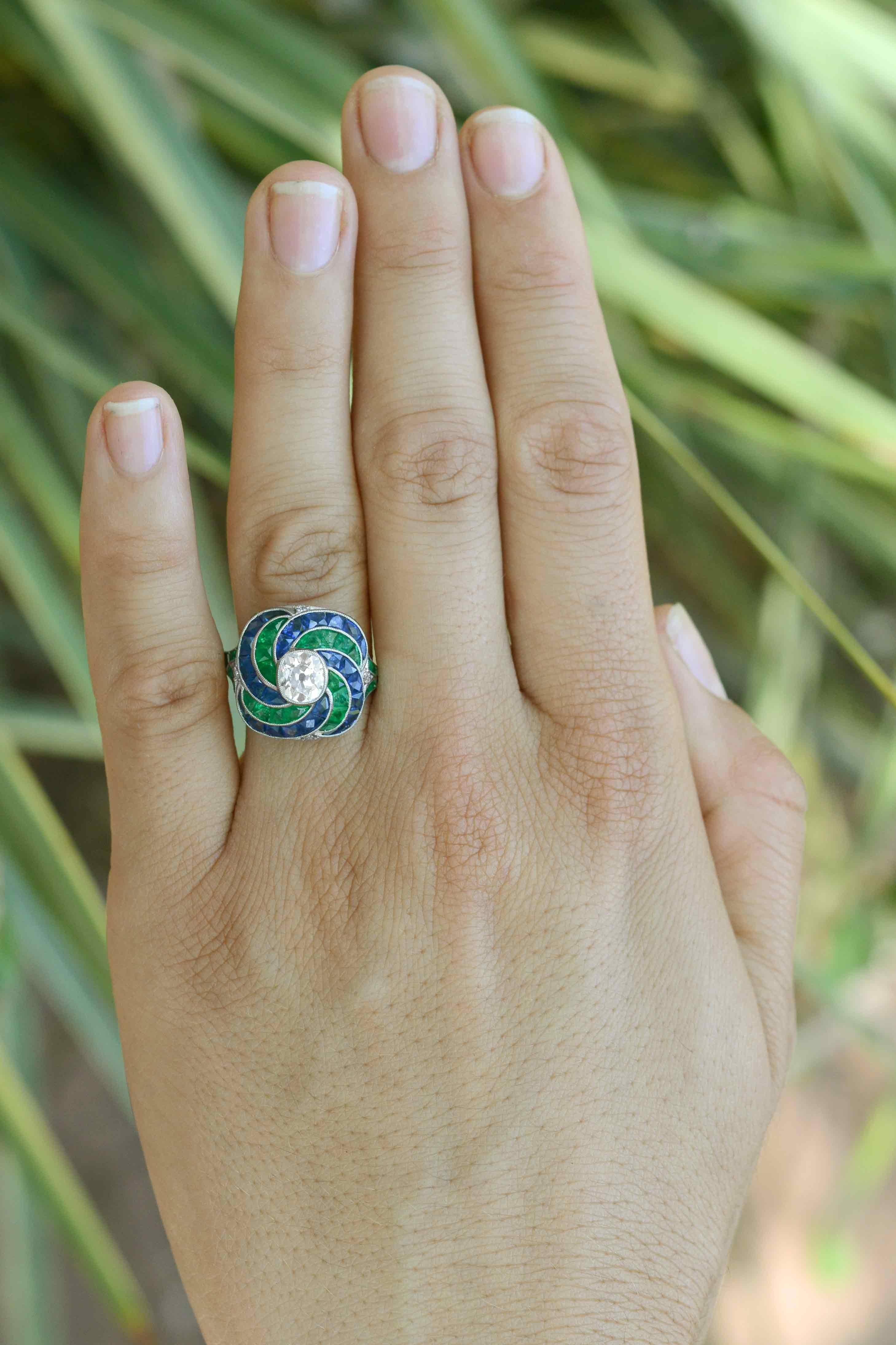A mesmerizing ensemble of diamond, emerald and sapphire, a swirling engagement ring. This Art Deco inspired, handmade platinum masterpiece, a faithful emulation of an antique Jazz Age 1920s heirloom centers upon an antique, old mine cut diamond near