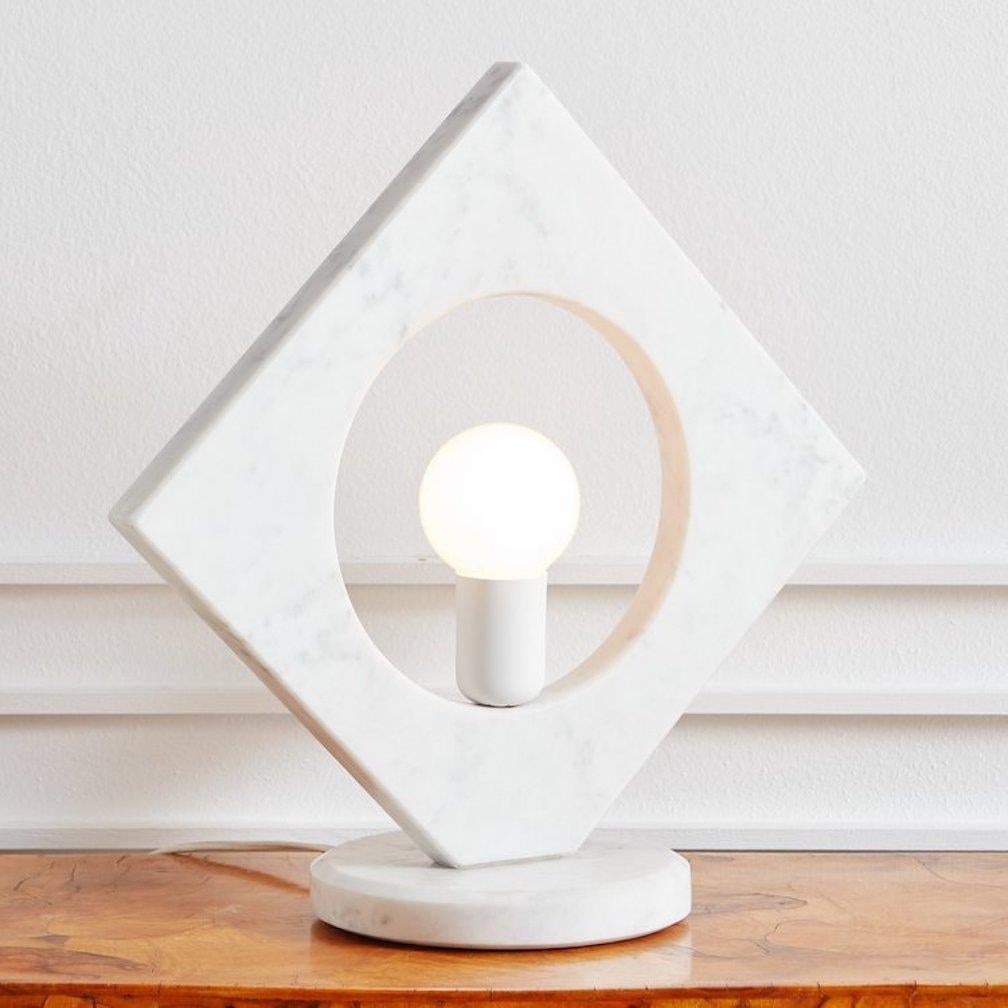 A handsome, geometric table lamp featuring beautiful Carrara marble with subtle gray veining. This lamp has a 1.25” thick angular marble body with a circular cutout that displays a single light bulb. It sits on an attached 1”H circular base with a