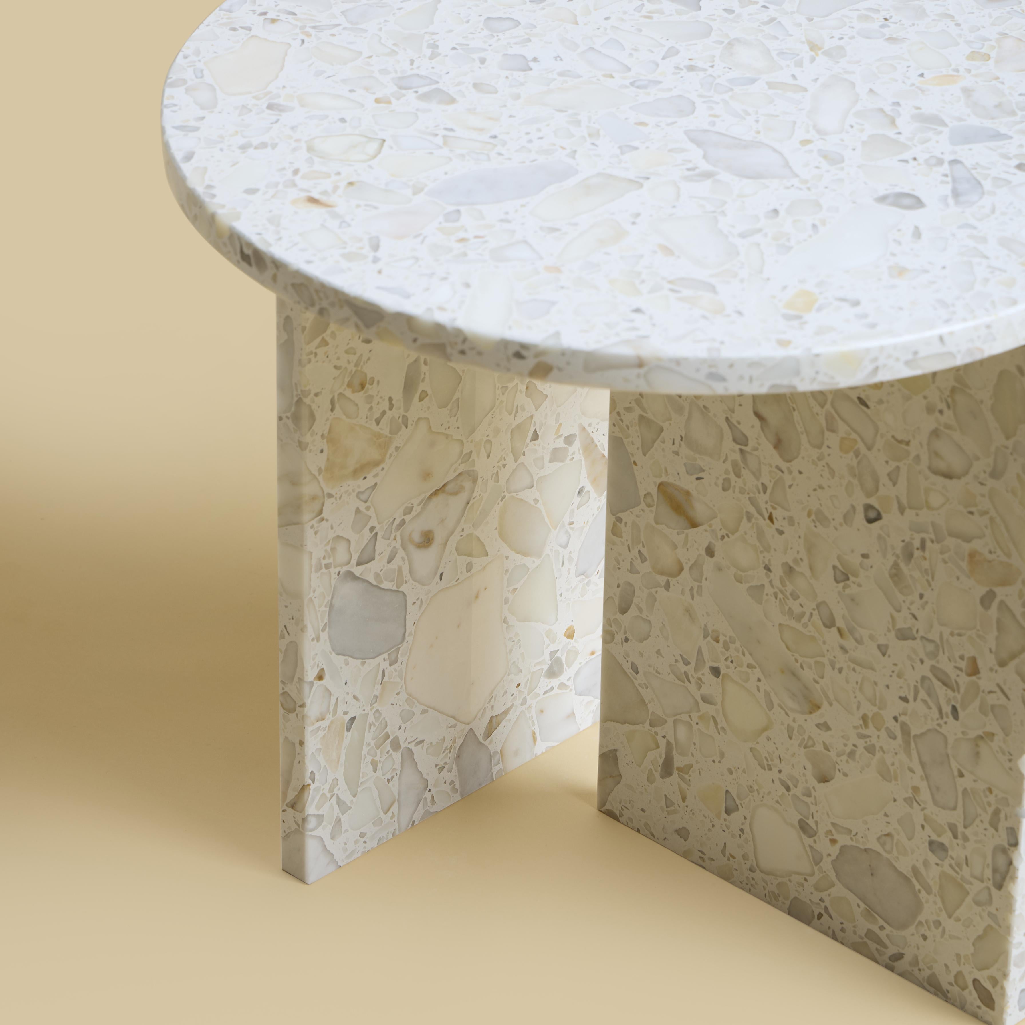 La table basse Kyushu est entièrement réalisée en Carrara Terrazzo. Le plateau est circulaire et mesure 45 cm de diamètre. Les pieds sont constitués de deux plaques de marbre dont une partie est incrustée sur le plateau, ce qui constitue un détail
