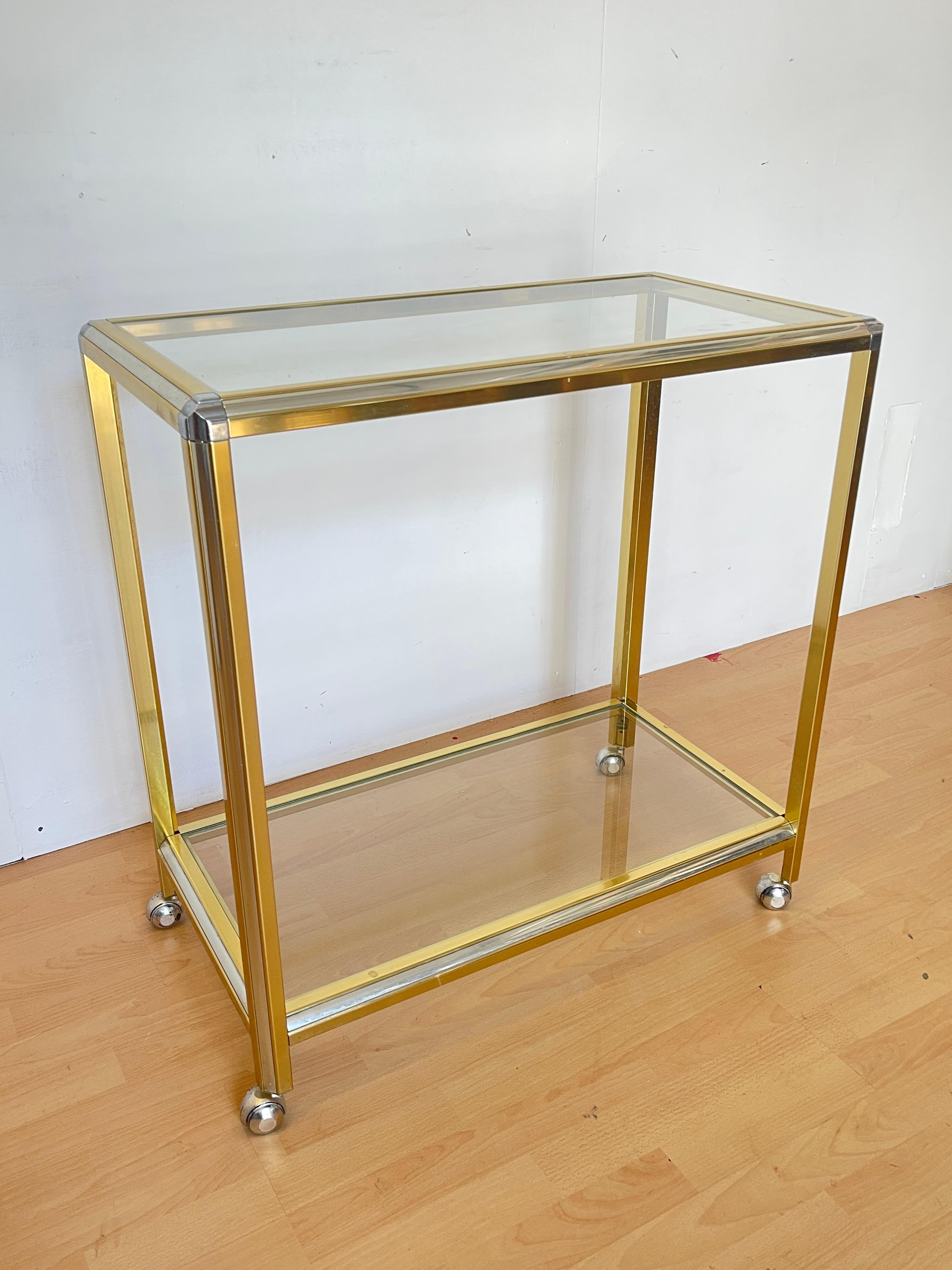 Cet élégant chariot de bar vintage, datant des années 1970, est un parfait exemple du design Hollywood Regency : essentiel dans sa forme mais luxueux dans ses matériaux.

La structure géométrique en métal doré brillant est associée à deux étagères