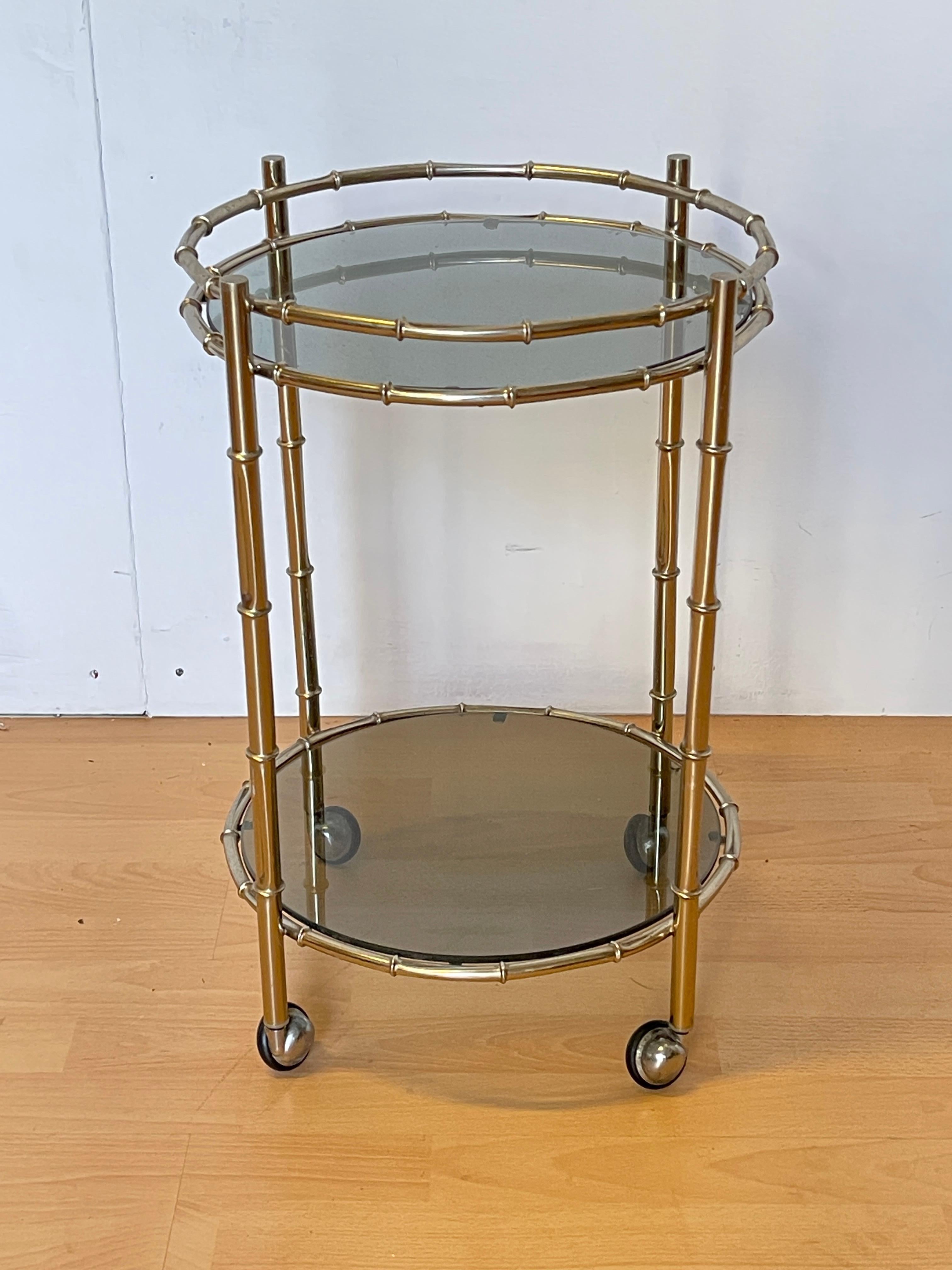 Chariot de bar élégant au design vintage, composé d'une structure métallique effet laiton travaillée en faux bambou. Équipé de deux étagères rondes en verre fumé, parfaites pour servir des boissons ou présenter des objets décoratifs. Les roulettes