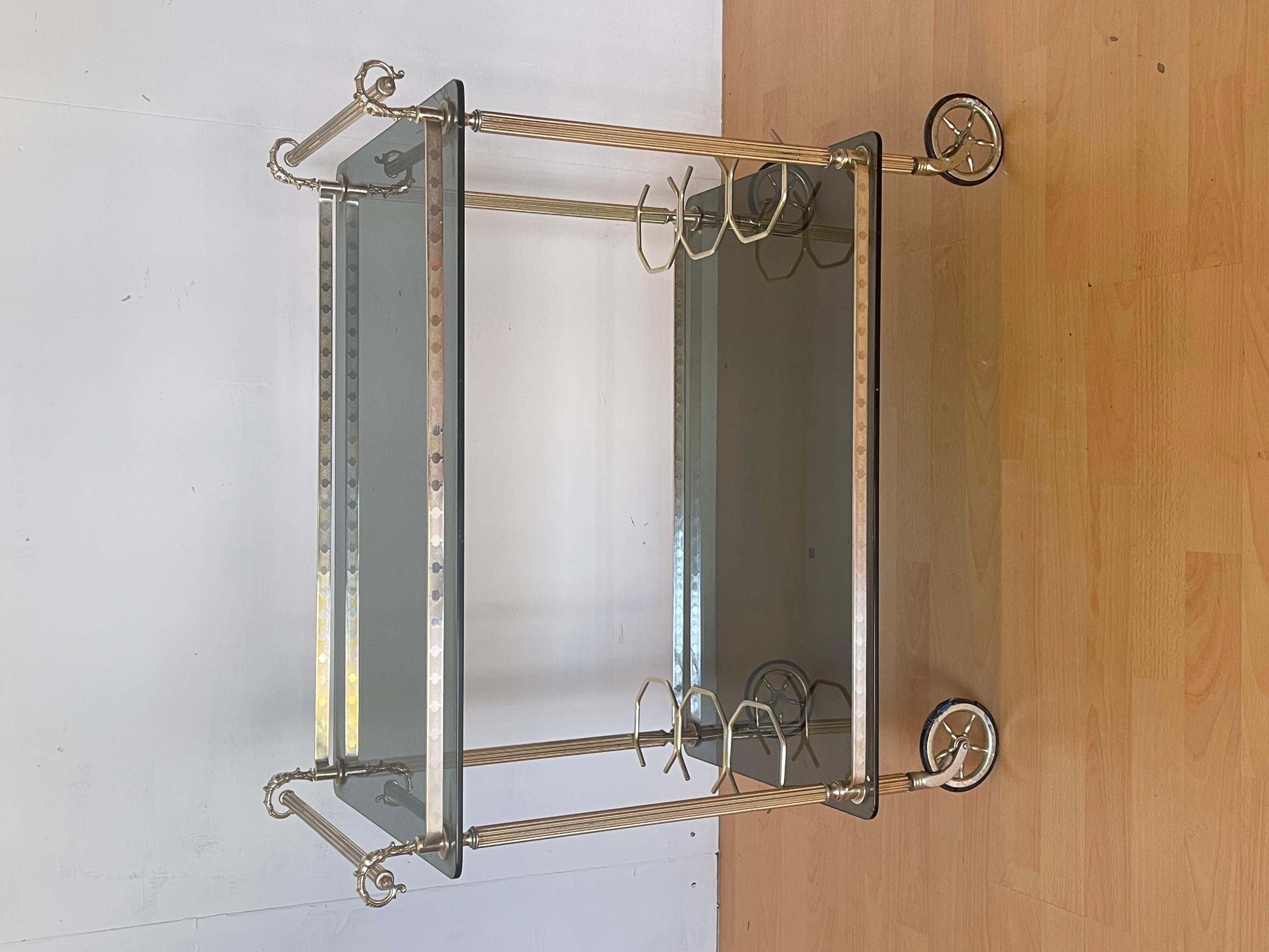 Elégant chariot de bar vintage des années 1960 au design raffiné et intemporel.
Le cadre en laiton chromé argenté présente une finition usinée avec des détails décoratifs rappelant le style Hollywood Regency, tandis que les deux étagères en verre