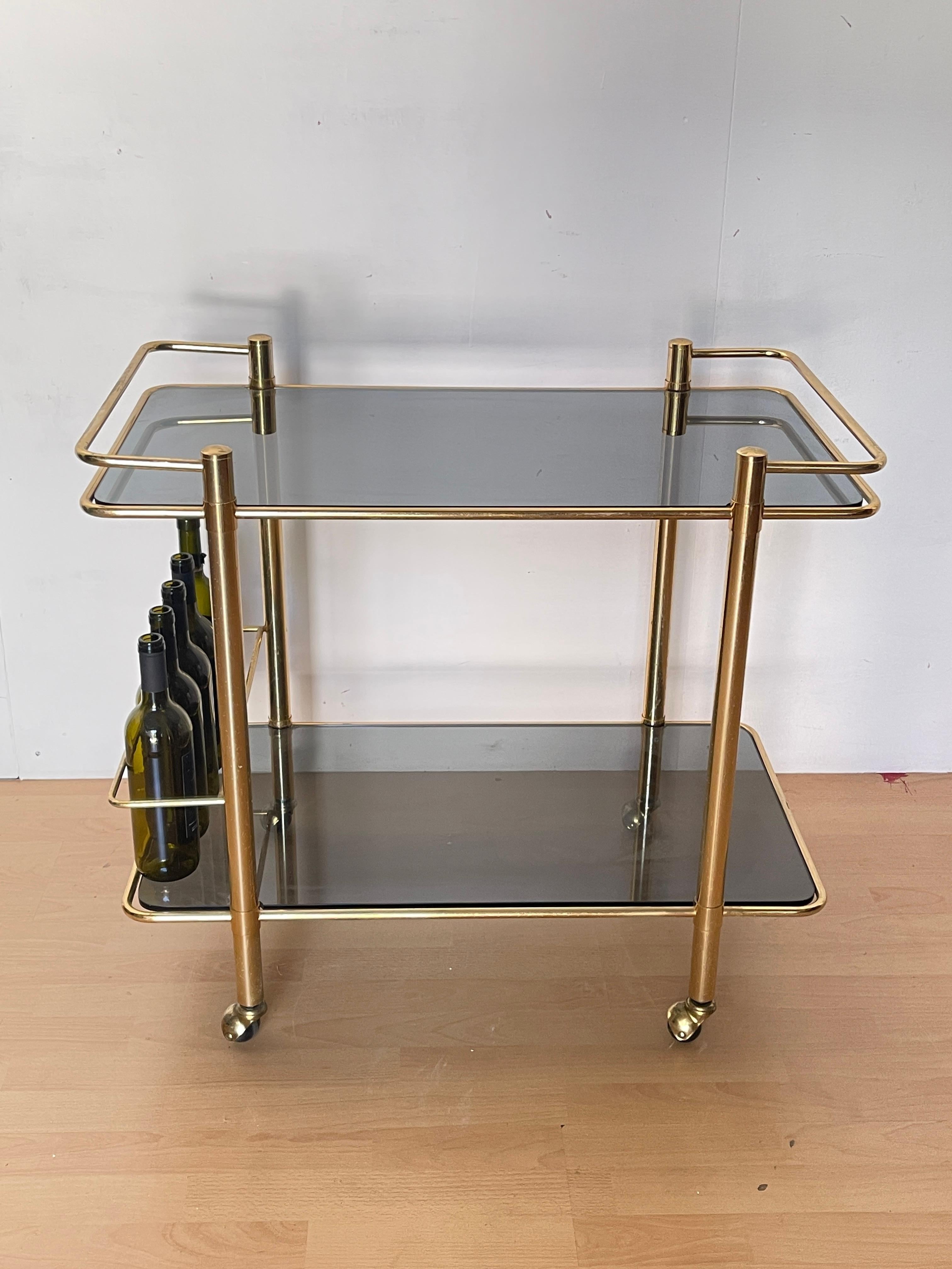 Affascinante carrello bar vintage in stile anni '70, perfetto per donare un tocco di eleganza retrò al tuo soggiorno, salotto o angolo cocktail.
La struttura è realizzata in otonne dorato, con ripiani in vetro fumé che donano un effetto raffinato e