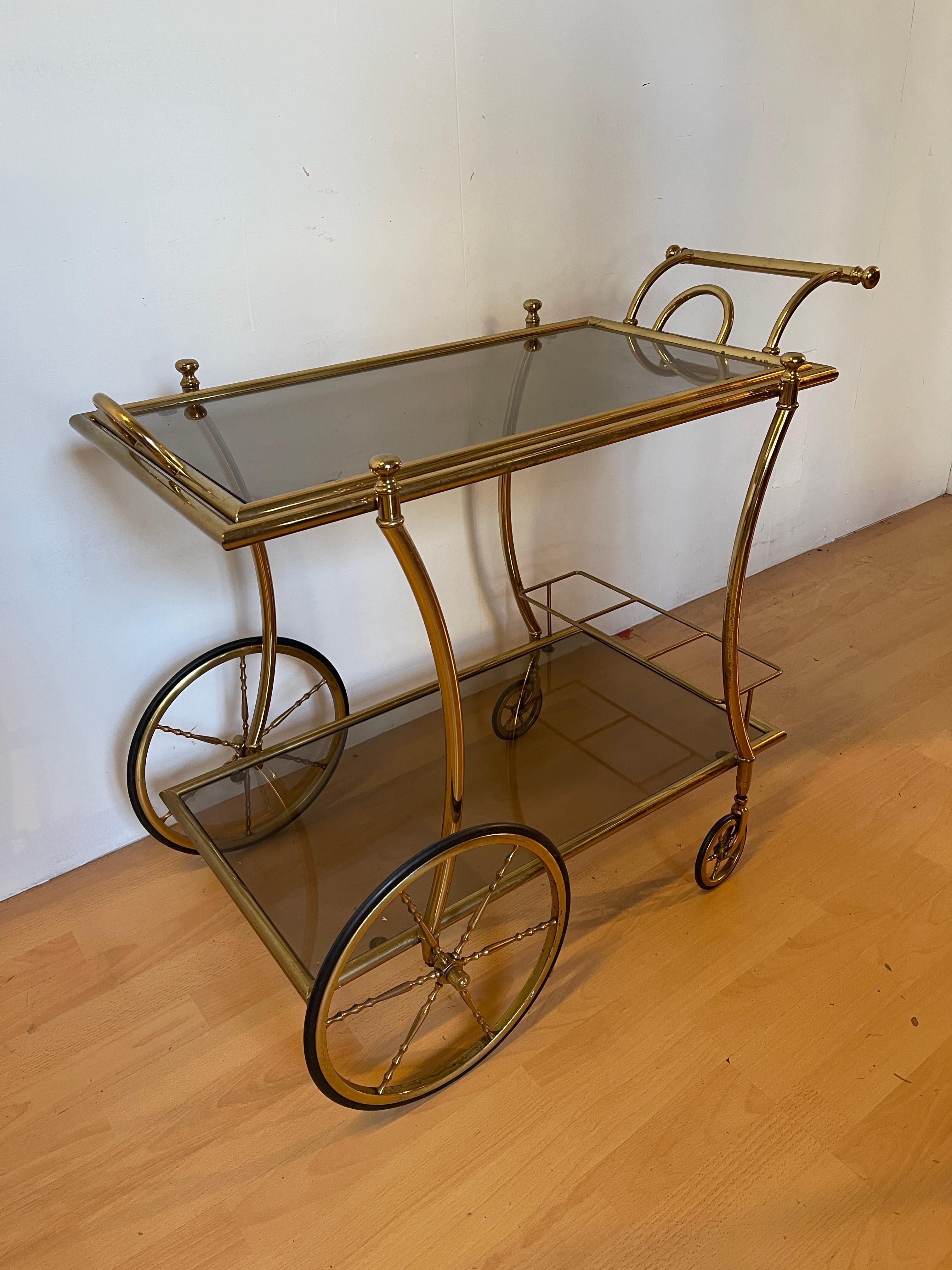 Elegante carrello da servizio o bar trolley in stile vintage, risalente agli anni ’60/’70. La struttura è realizzata in ottone dorato con linee curve e raffinate, mentre i due ripiani sono in vetro fumé, perfetti per esporre bottiglie, bicchieri o