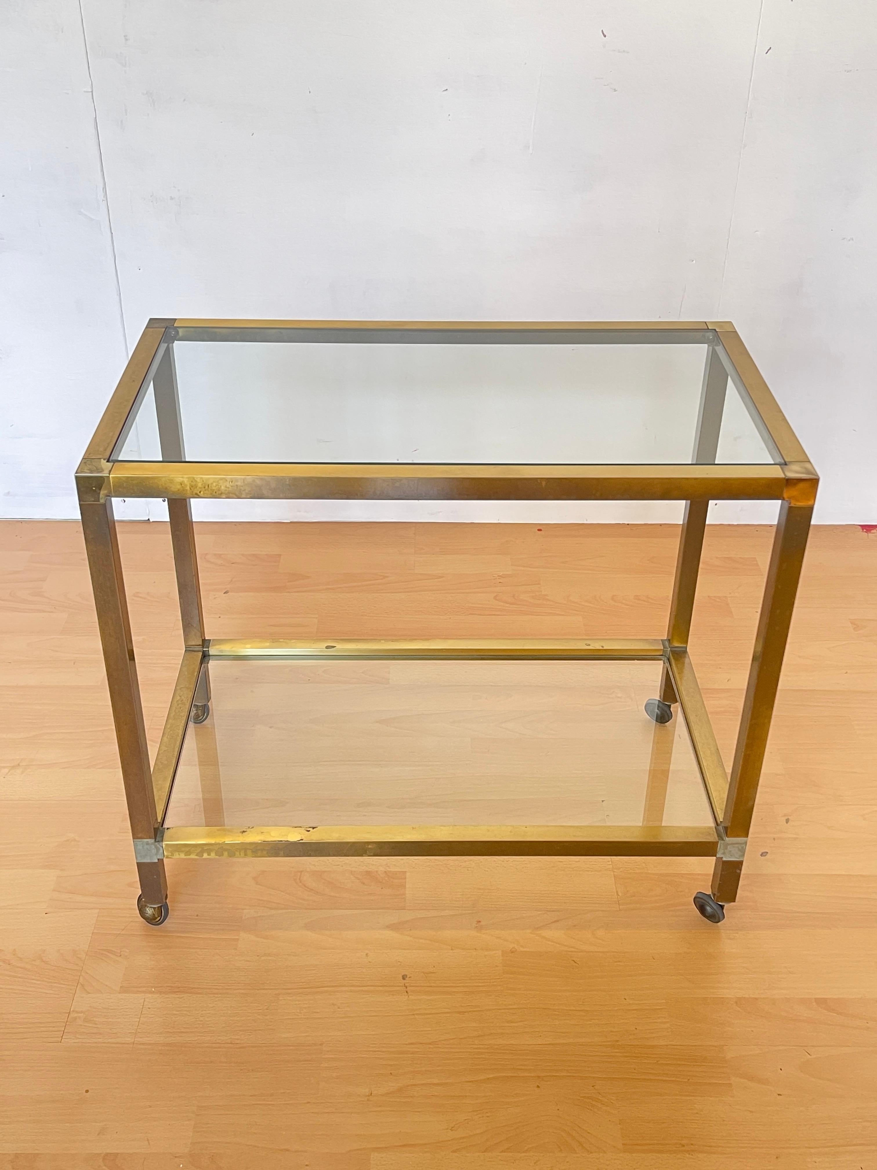 Elegante carrello da tè o da servizio in stile vintage, risalente agli anni ’70, con struttura in metallo ottonato e ripiani in vetro trasparente. Le linee pulite e geometriche, unite al fascino dell’ottone brunito dal tempo, donano a questo pezzo
