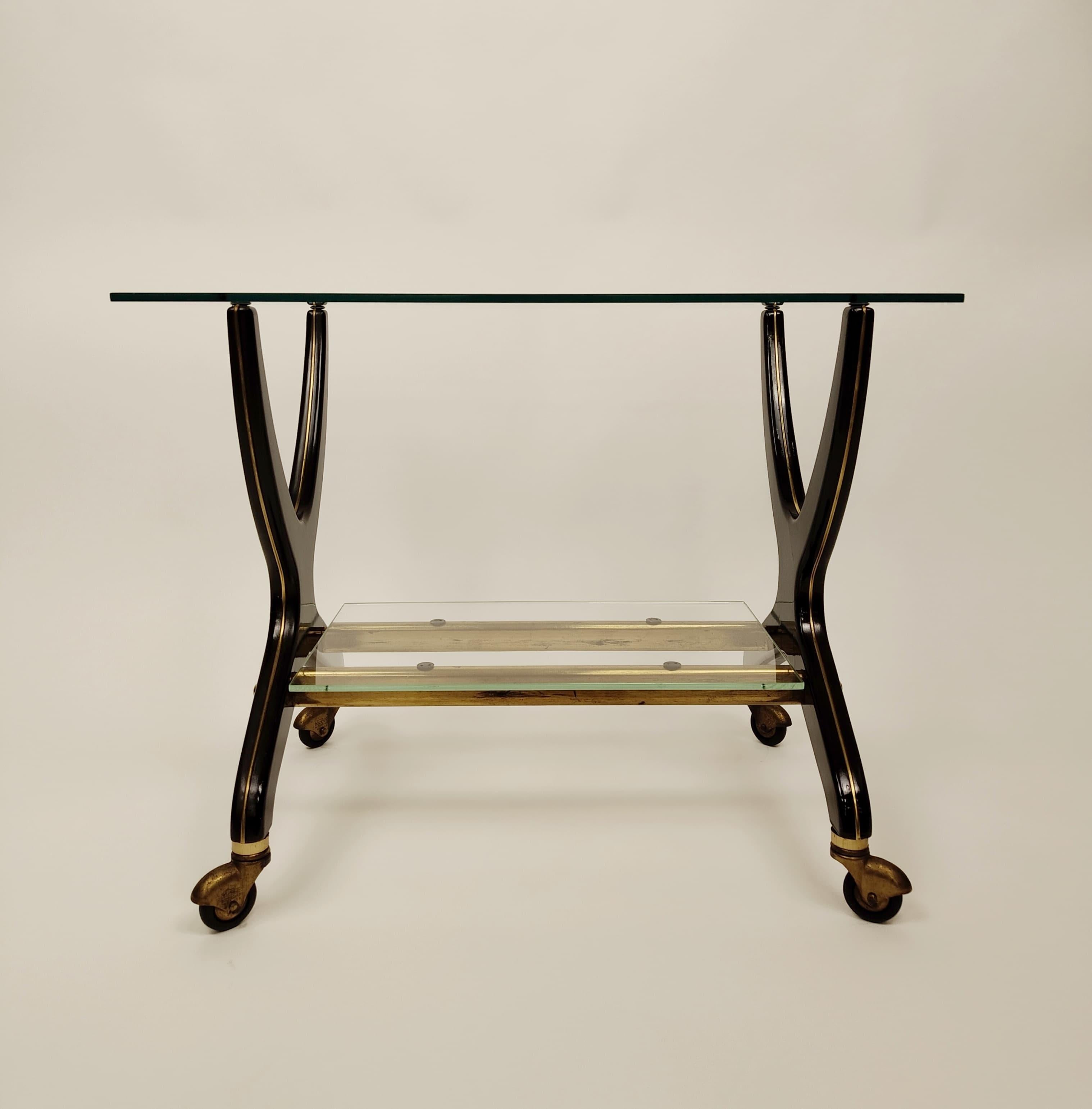 A cart  dating from about the 1950s measuring 48x60x42 cm.
The frame features curved wooden legs with a dark finish, embellished with gold details. The top and bottom surfaces are made of clear glass and offer a sense of lightness and modernity that