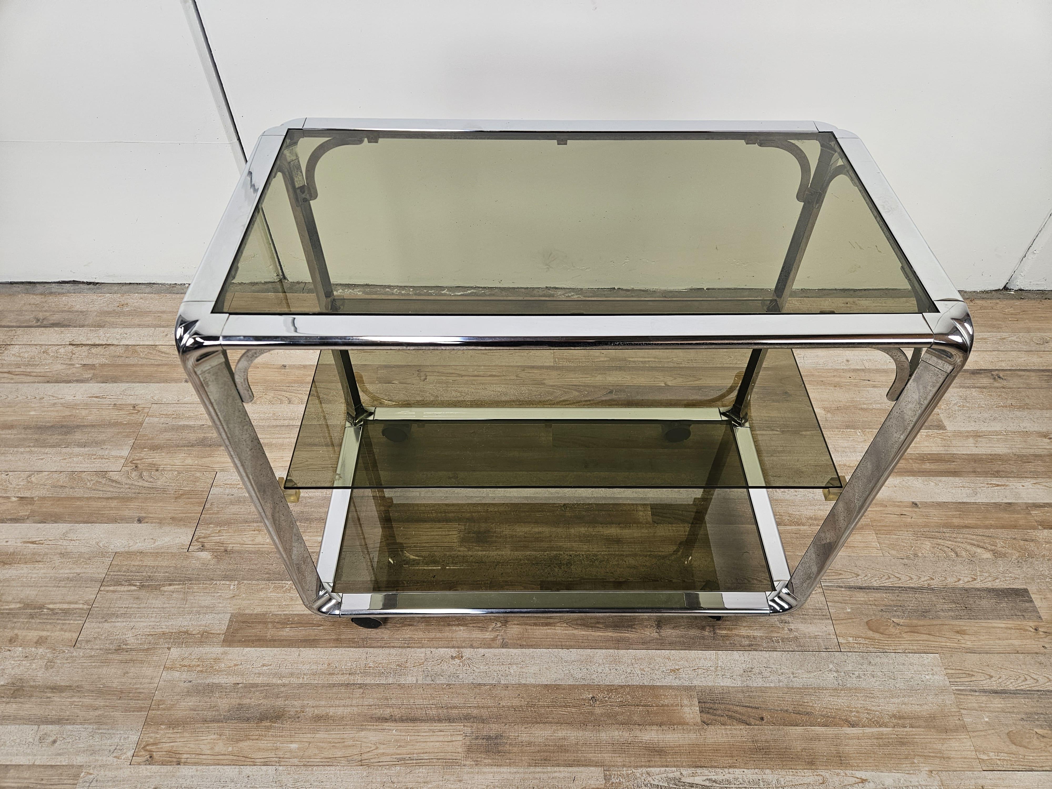 Carrello dei primi anni '70 italiano in stile Hollywood Regency, dotato di tre ripiani rettangolari in vetro fumè e rotelle alla base.

Altezza da terra del primo ripiano 8cm.
Altezza da terra del secondo ripiano 41cm.

Normali segni d'usura dovuti