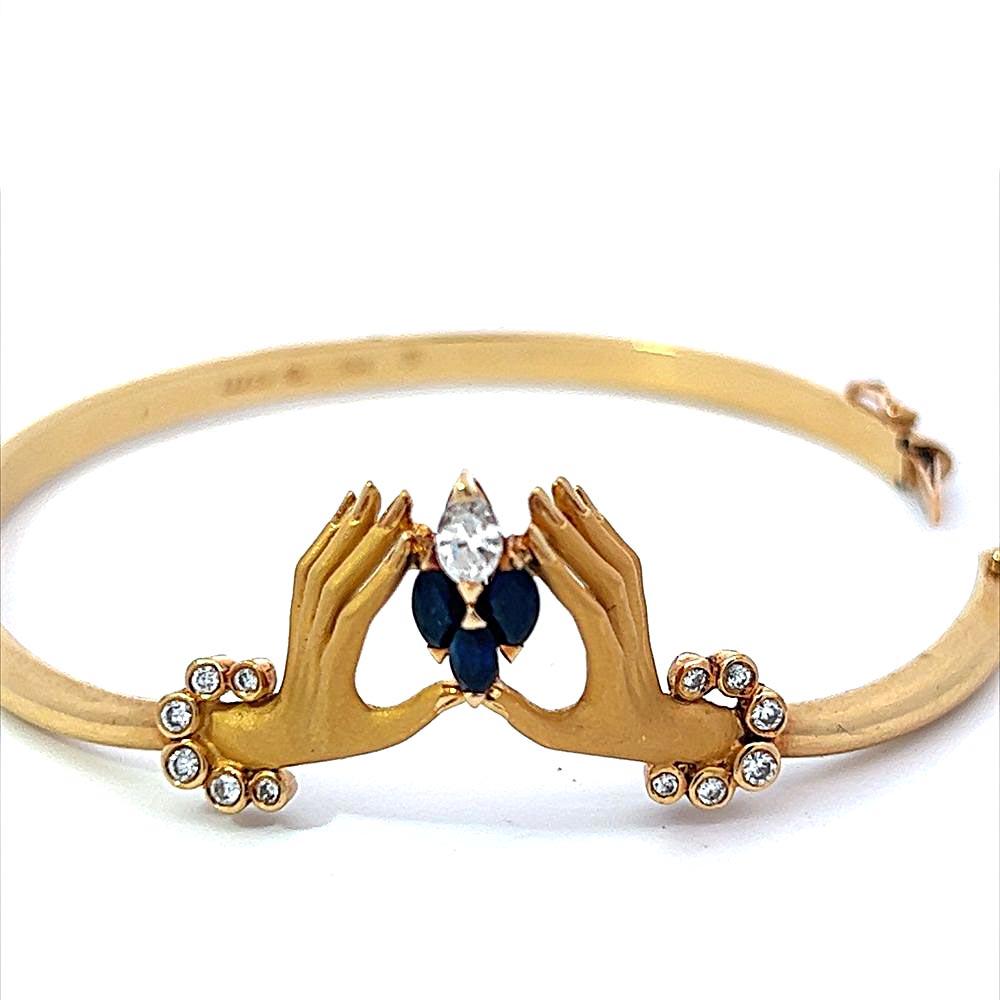 This exceptionally crafted Carrera y Carrera 18 karat yellow gold bracelet bangle features two beautifully detailed hands holding two blue sapphire marquise gemstones and one marquise shape white diamond. A small diamond bracelet accents each hand