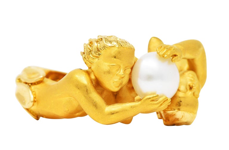 Ring is designed as two highly rendered matte gold cherubs with texturous hair

With hands extended to hold a 6.0 mm round pearl

White in body color with moderate iridescence and good luster

Accented by tulip motif shoulders and high polished