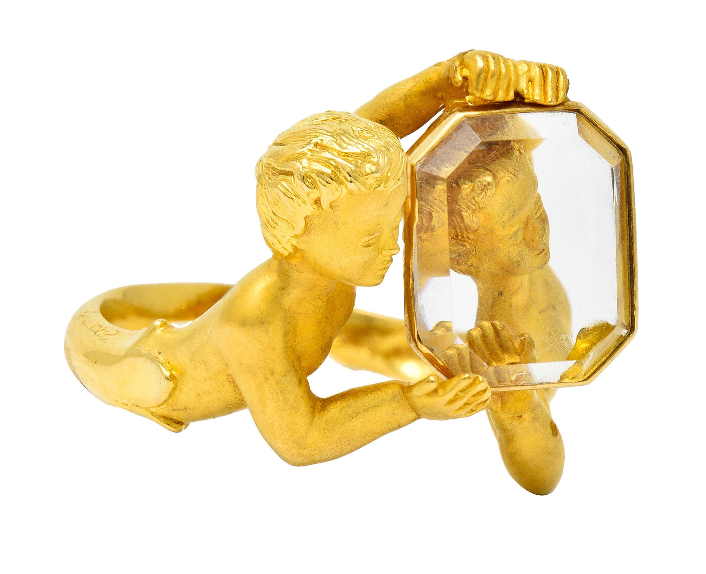 Bypass style ring is designed as two matte gold and highly rendered cherubs

Holding and playfully peering through an emerald cut tablet of crystal

Transparent with and excellent polish while measuring approximately 12.0 x 10.0 mm

Completed by a