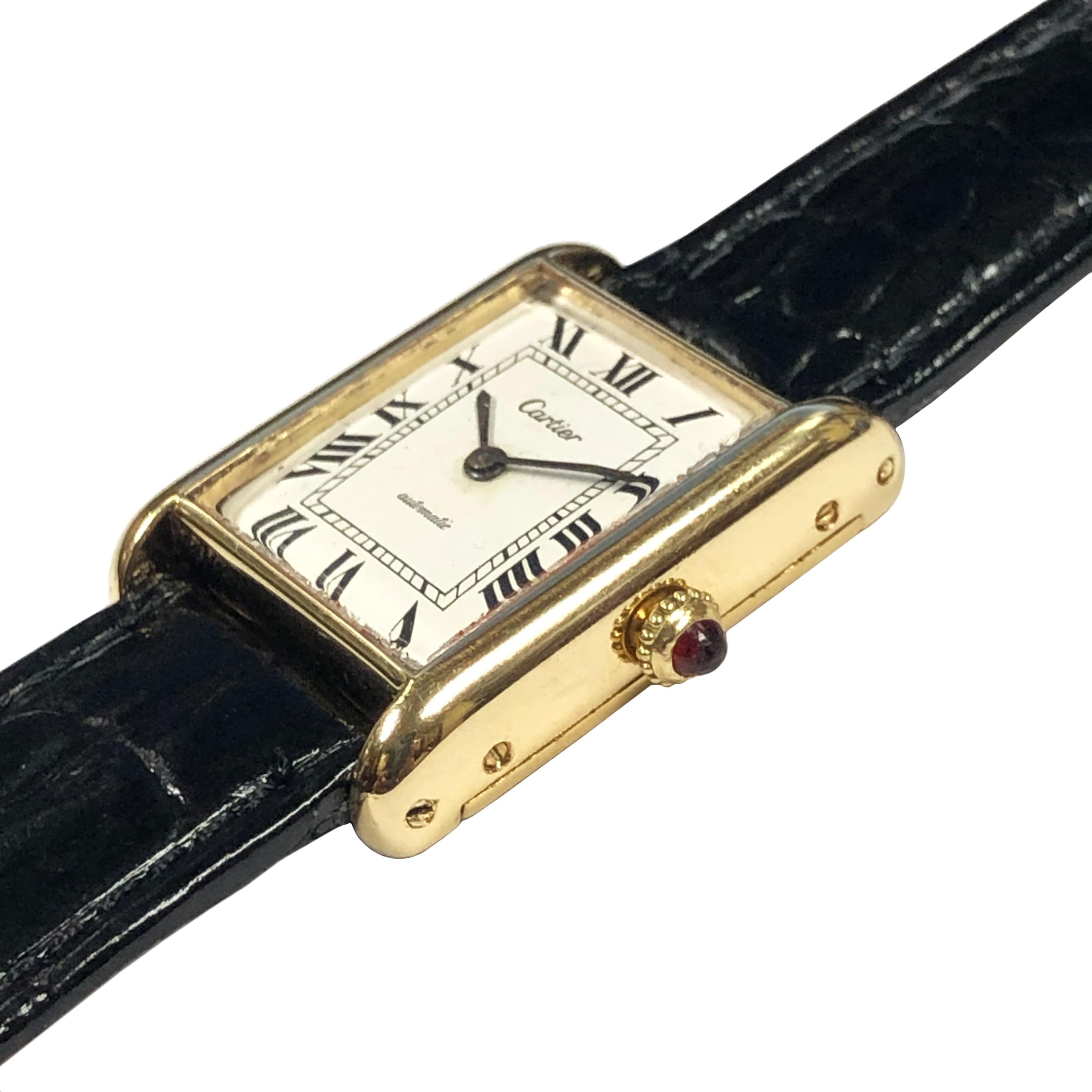Circa 1980 Cartier Classic Tank Wrist Watch, 30 X 23 M.M. 18K Yellow Gold 2 Piece case, Automatic, self winding movement, White dial with Black Roman Numerals, Ruby crown. New Hadley Roma Black leather strap with a Cartier Gold Plate Tang Buckle.