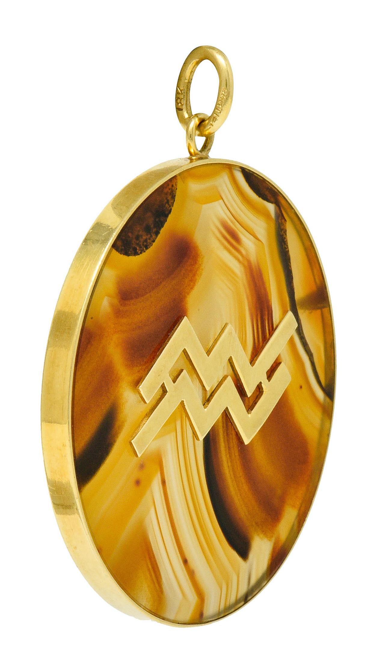 Large circular pendant features a bezel set tablet of agate

Semi-transparent and colorless with brown to yellow ochre banding throughout

Centering a highly polished gold emblem depicting stylized zodiac waves of Aquarius

Completed by a jump ring
