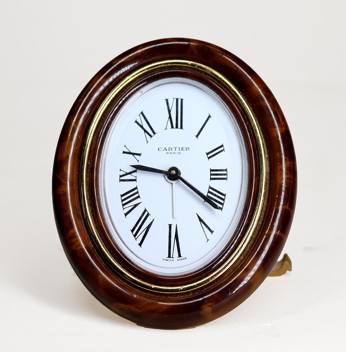 A Cartier Les Must Alarm Clock in an oval enamel and gold plated frame. The enamel giving the appearance of polished burr walnut, with cabochon jewelled setting nuts, resting on a strut back with the distinctive Cartier ‘C’ forming the feet.

The