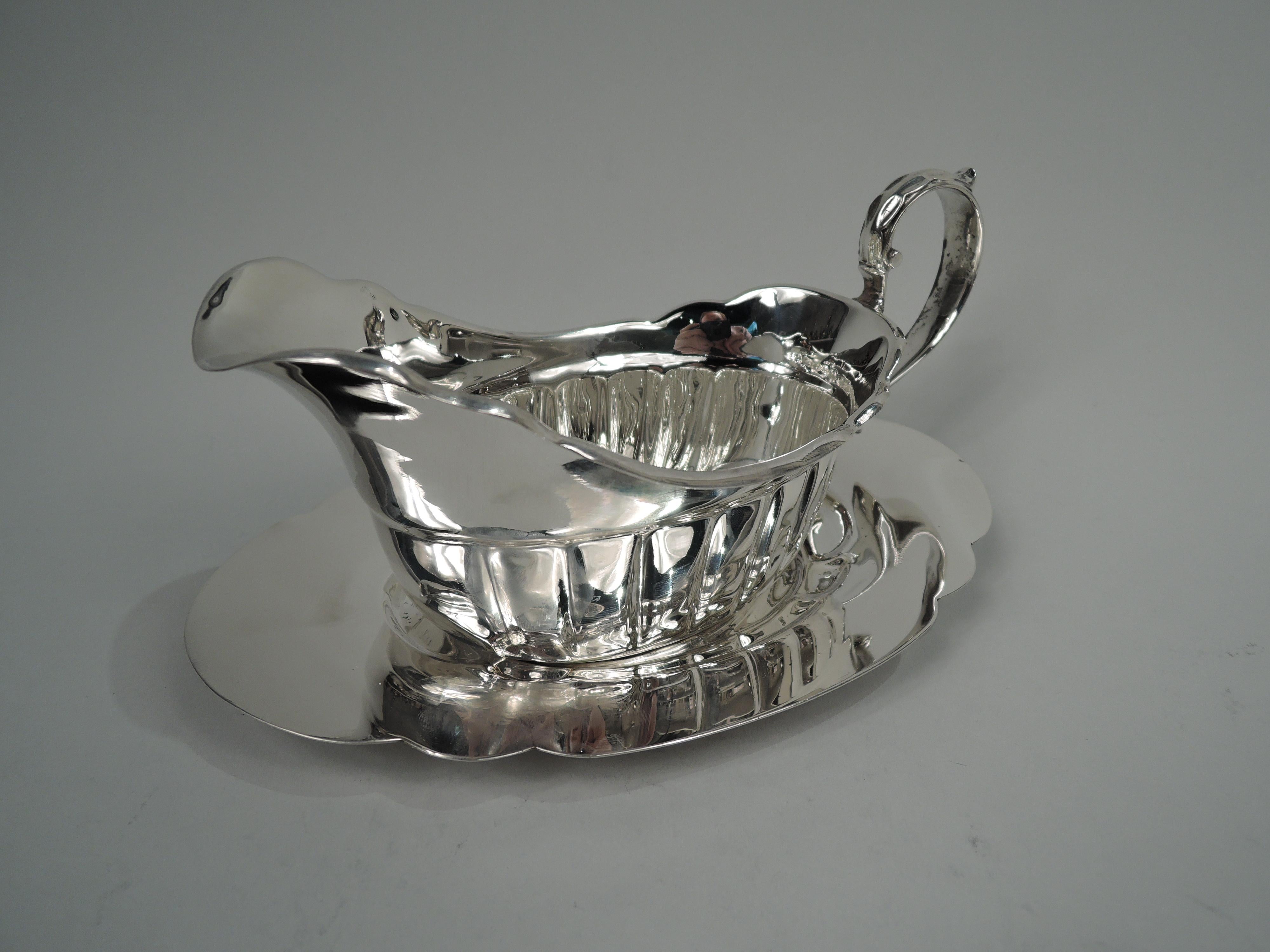 American Classical sterling silver gravy boat on stand, ca 1950. Retailed by Cartier in New York. Boat: Oval lobed bowl with u-form spout and leaf-capped high-looping double-scroll handle. Stand: Oval well in shaped scrolled surround with wide