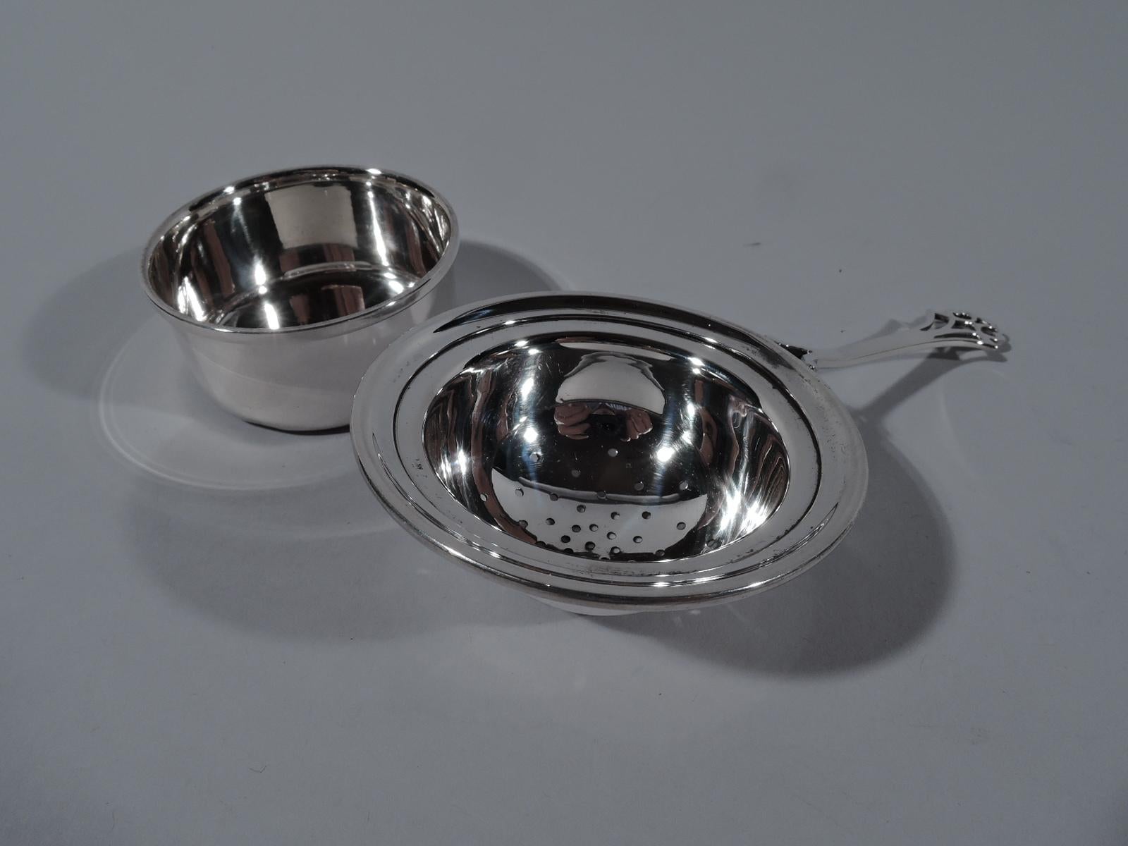 American modern sterling silver tea strainer on stand. Retailed by Cartier in New York, circa 1930. Strainer has pierced round bowl with molded rim. Split-mounted tapering handle with curvilinear open terminal. Stand round with straight sides and