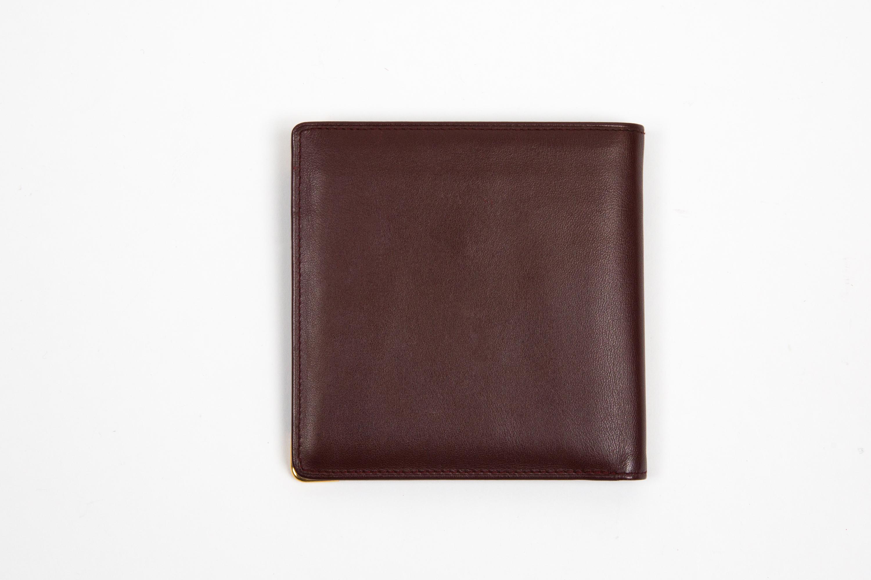 Viereckige Brieftasche aus bordeauxfarbenem Leder von Cartier mit quadratischer Form, vergoldeten Ecken, Innenfächern für Karten und Geld und einer goldfarbenen Innenmarke. 
Circa: 1990er Jahre
In gutem Vintage-Zustand. Hergestellt in Frankreich.
11