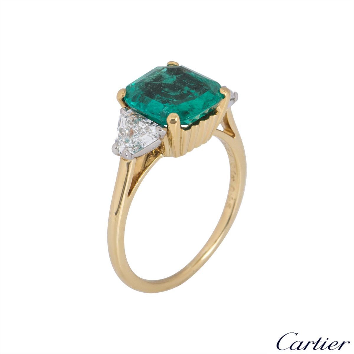 An 18k yellow gold emerald and diamond ring by Cartier. The ring comprises of a cushion cut emerald set with a single trillion diamond on each side in a platinum claw. The cushion cut emerald has an approximate weight of 2.26ct and the trillion cut