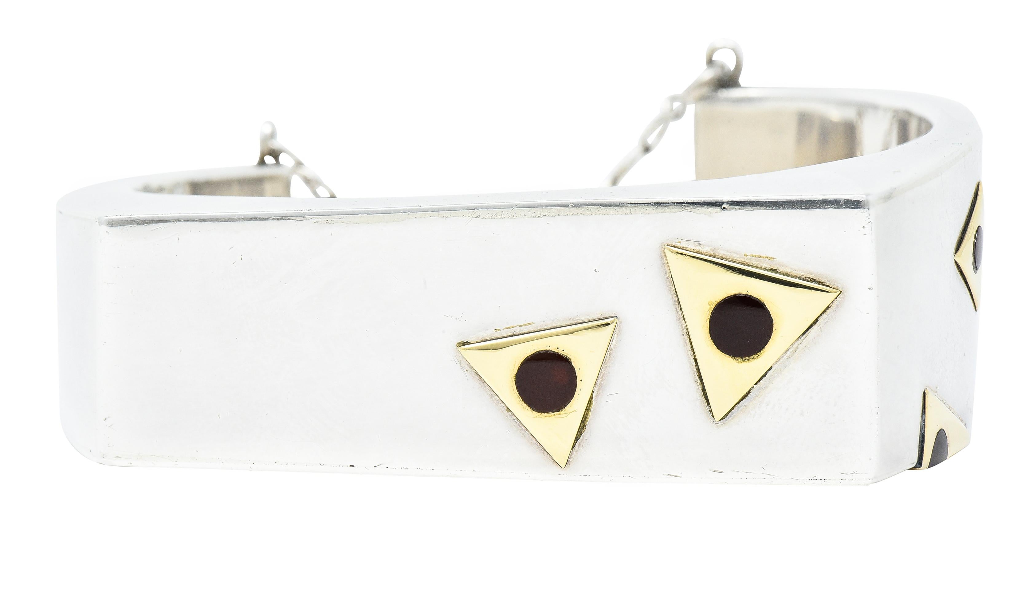 Disegnato come un bracciale asimmetrico in argento sterling con la parte superiore squadrata. Caratterizzato da un motivo a triangolo in oro giallo su un lato. Al centro cerchi di corniola intarsiati di dimensioni variabili. Colore rosso arancio