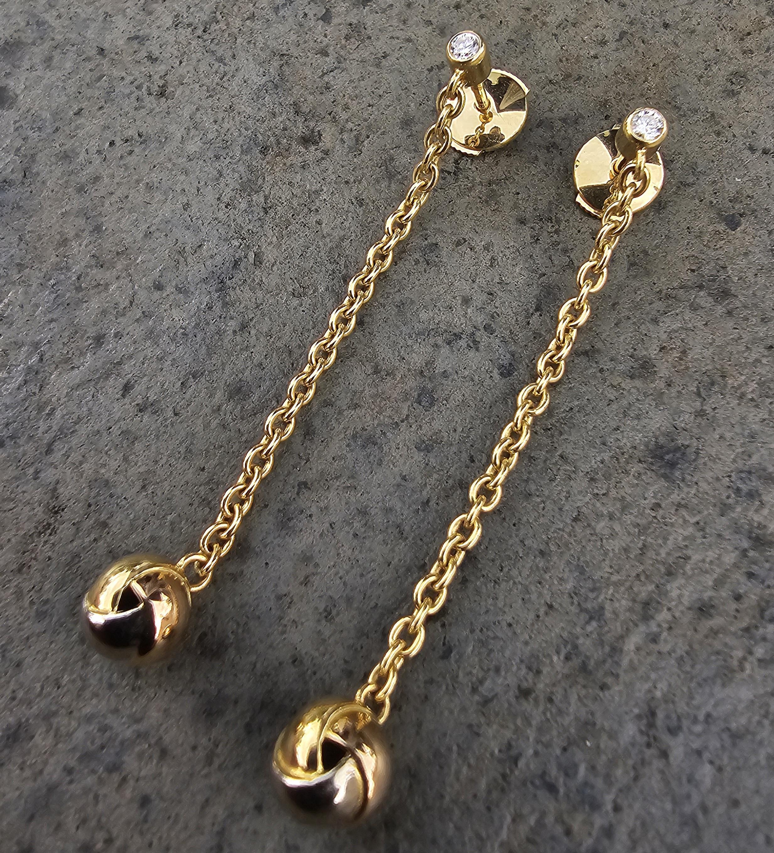 Estos pendientes colgantes Cartier muestran un diseño refinado en oro amarillo de 18 quilates. Los pendientes presentan una delicada cadena que suspende con elegancia un dije esférico de Trinidad, añadiendo un grácil movimiento. En la parte