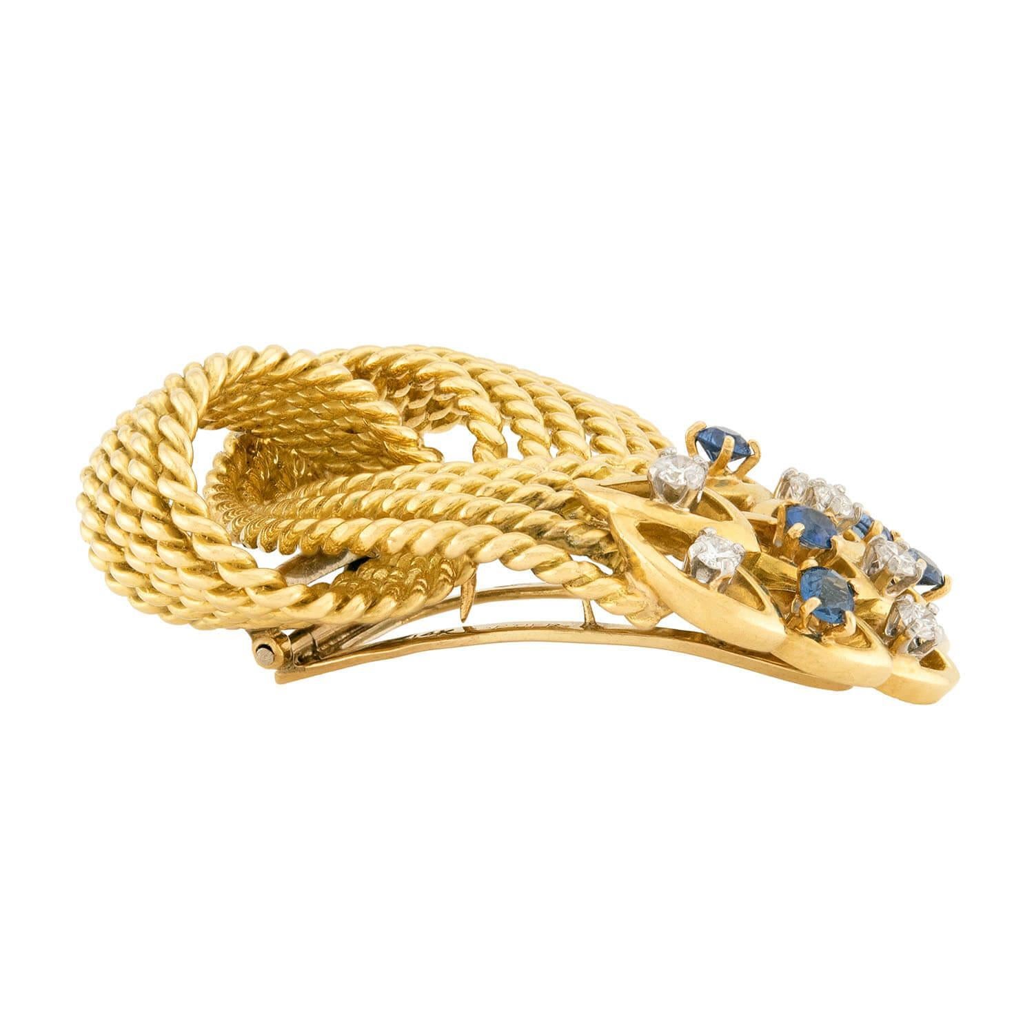 An absolutely exquisite Cartier diamond and sapphire pin! Crafted in vibrant 14k yellow gold, this substantial pin features a large interlocking knot-motif, which is composed of thin, gold-braided components. The pin also features 6 modern round