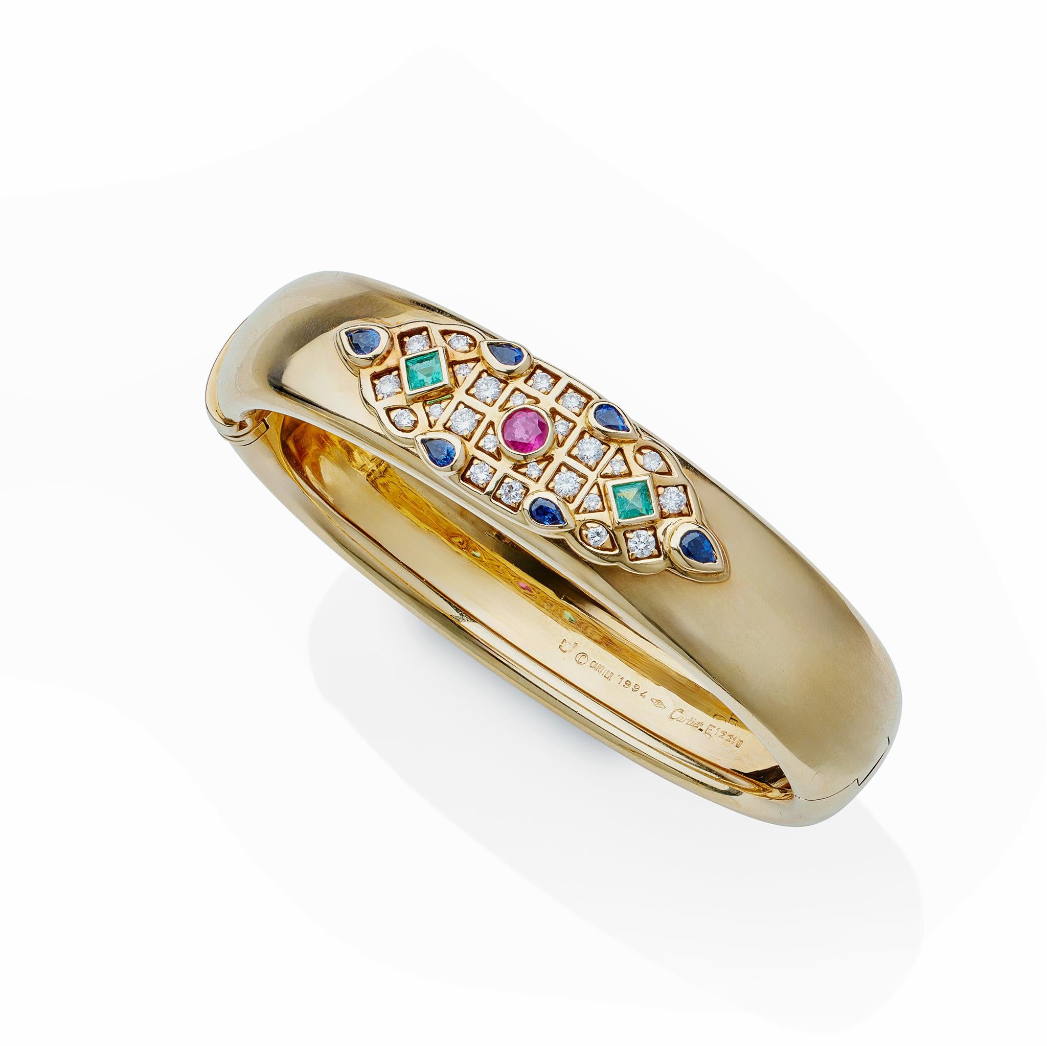 Created in 1994, this 18K gold, ruby, emerald, sapphire and diamond bangle bracelet is a jewel by Cartier. It is designed as a hinged bangle, its top set with an applied design centering a circular-cut ruby, six pear-shaped sapphires, and two