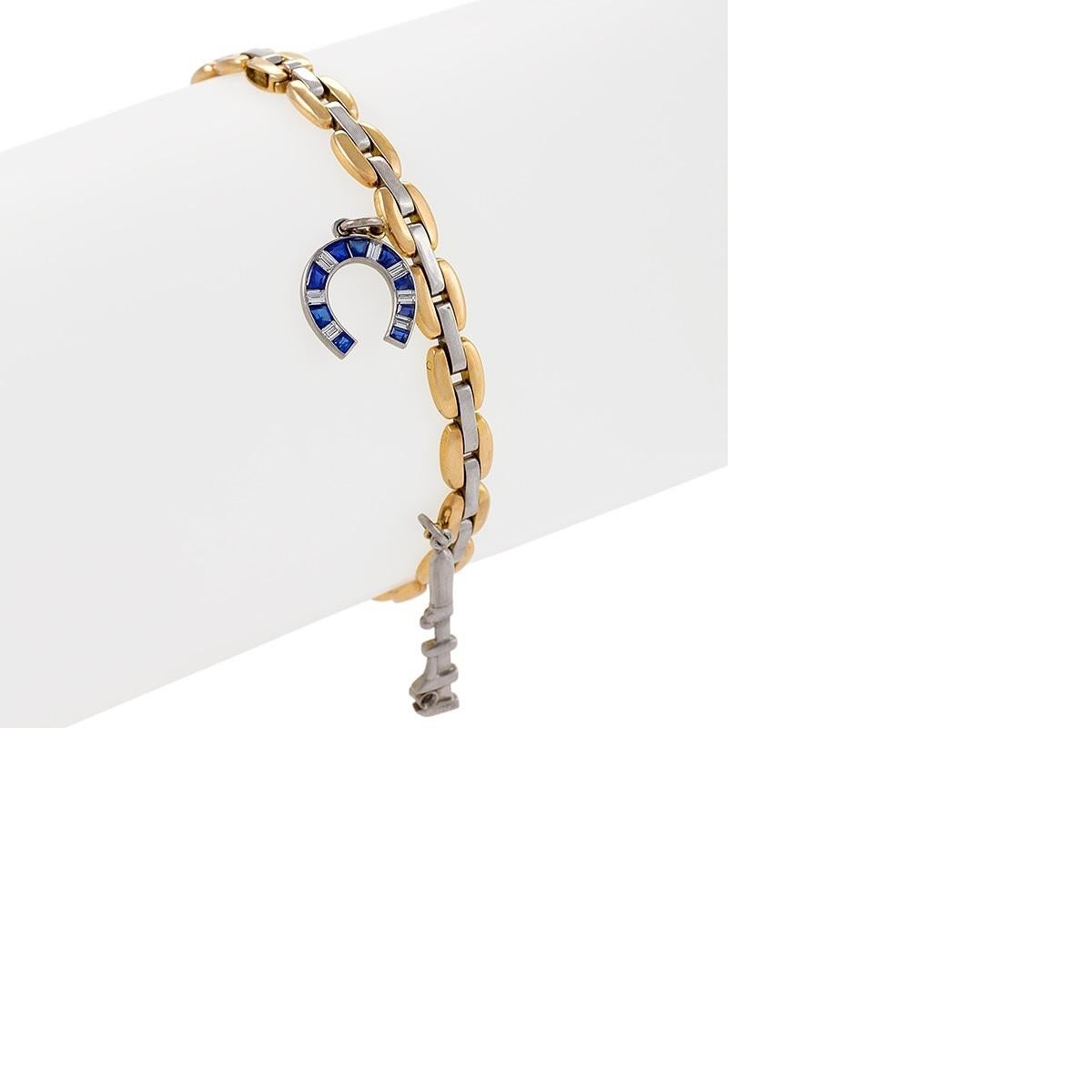 A two tone bracelet in platinum and 18k yellow gold with charms by Cartier. The bracelet has five charms, a sapphire and diamond horseshoe, a diamond accented wrench, a pair of fully articulated scissors, a house with emerald and sapphire accents