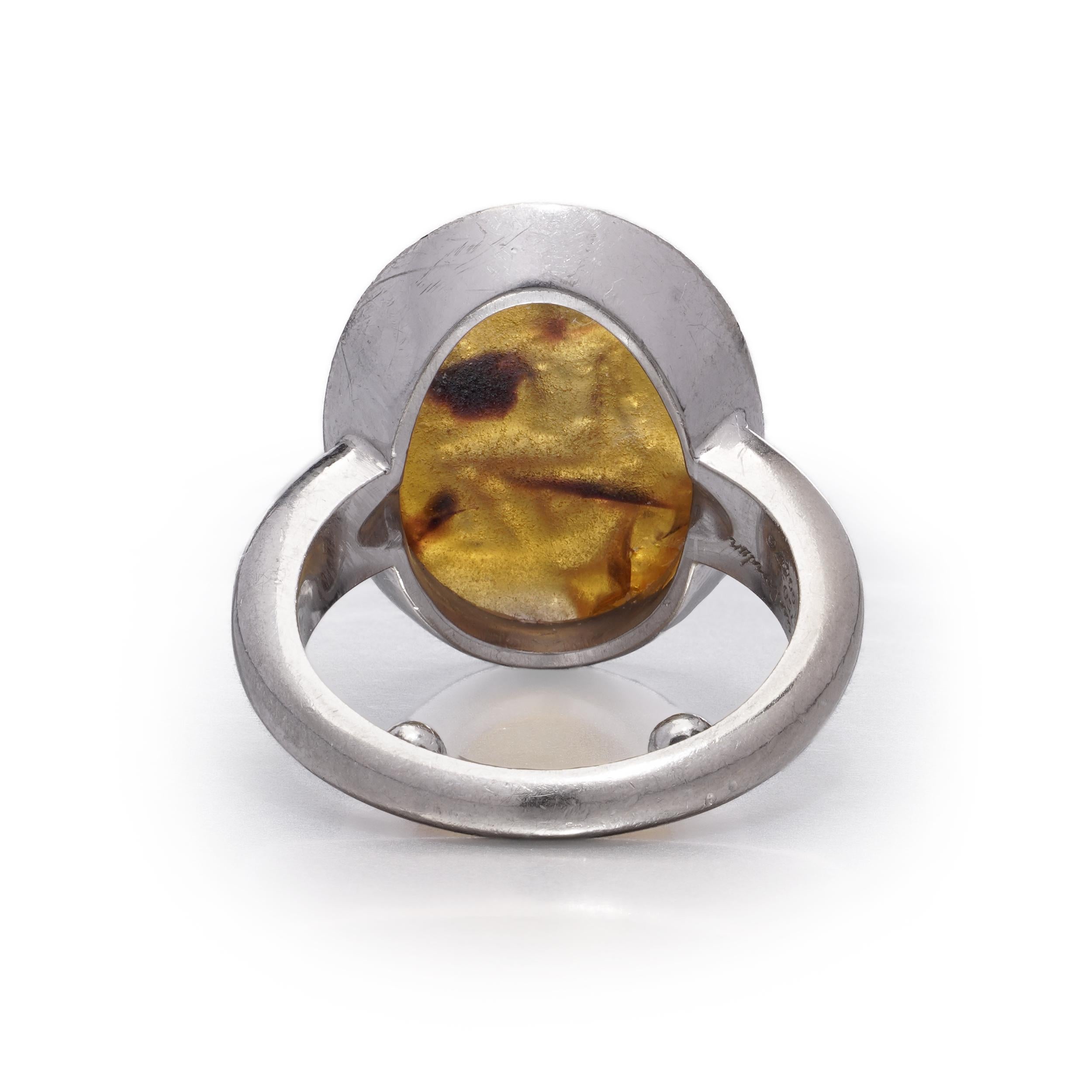 A bold and sculptural vintage ring by mounted by Cartier, crafted in 18kt white gold and bezel-set with an oval amber cabochon. 
The amber intaglio carved to depict a seated figure.
Signed Mtd. Cartier London, maker's mark J.C, numbered, London