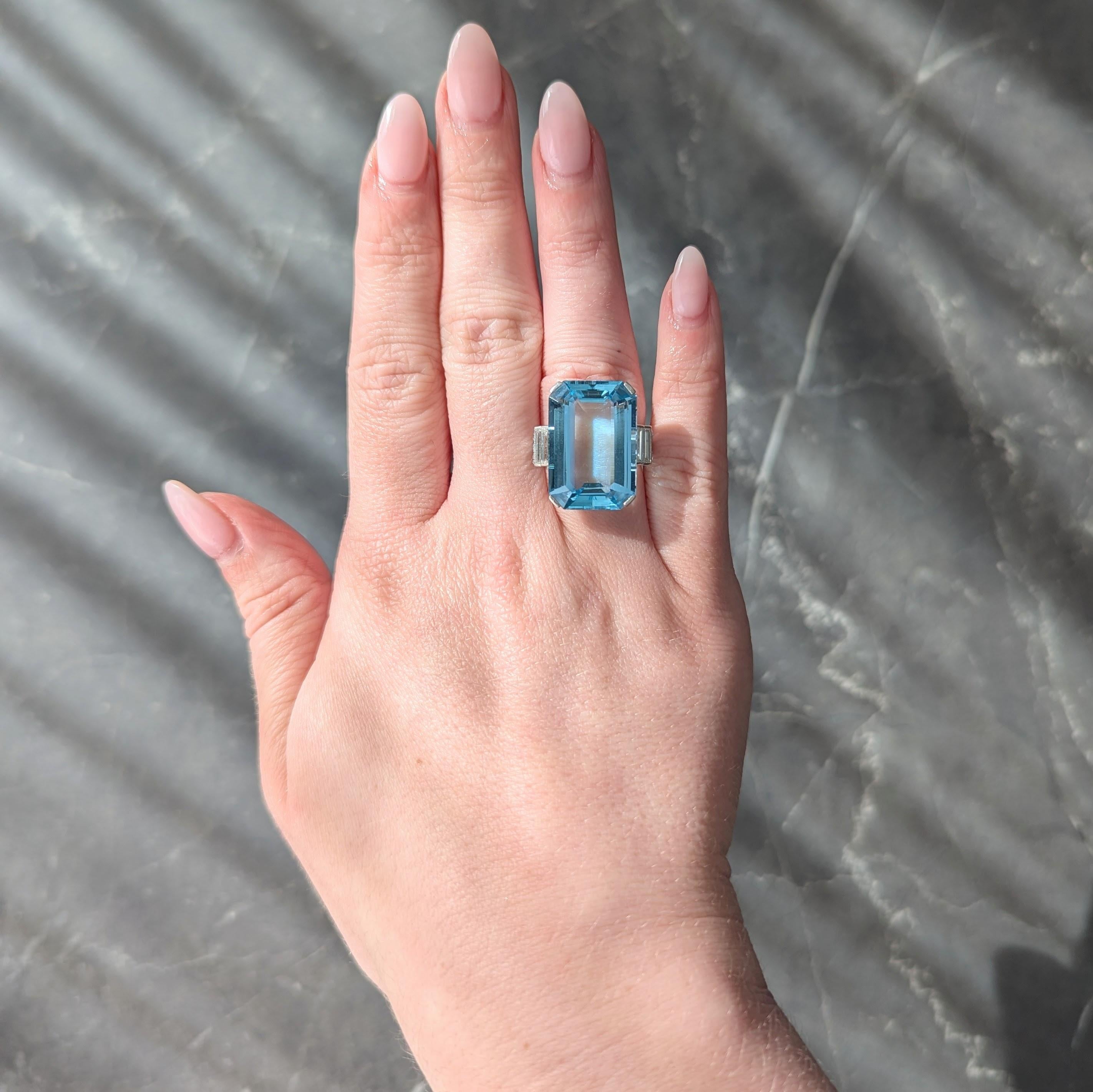 Centering an emerald cut aquamarine weighing approximately 21.24 carats
Transparent light blue in color and set in basket via corner tab prongs
With open wire gallery and flanked by geometric cathedral shoulders
Bezel set with baguette and round