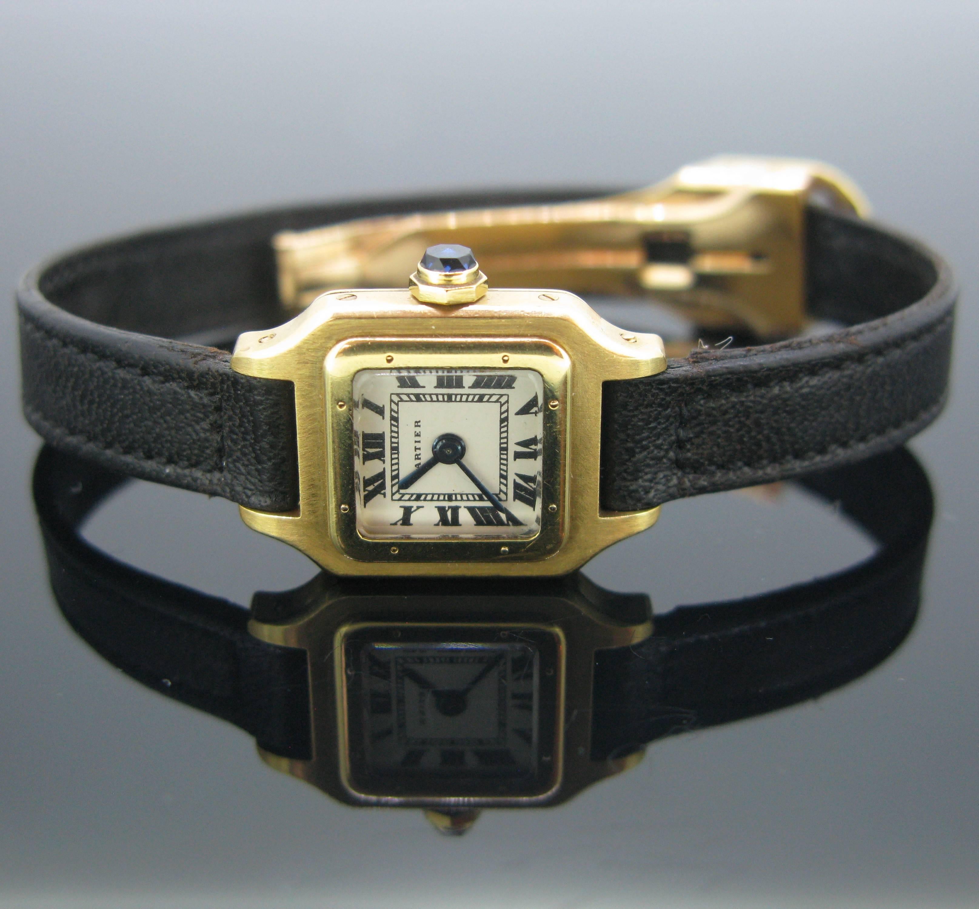 Cartier Mini Santos Manual Winding wrist watch. The dial is white with black Roman numerals. The square case is made in 18k Yellow Gold and the beaded crown is set with a synthetic faceted sapphire. The strap is in black leather and with an 18k