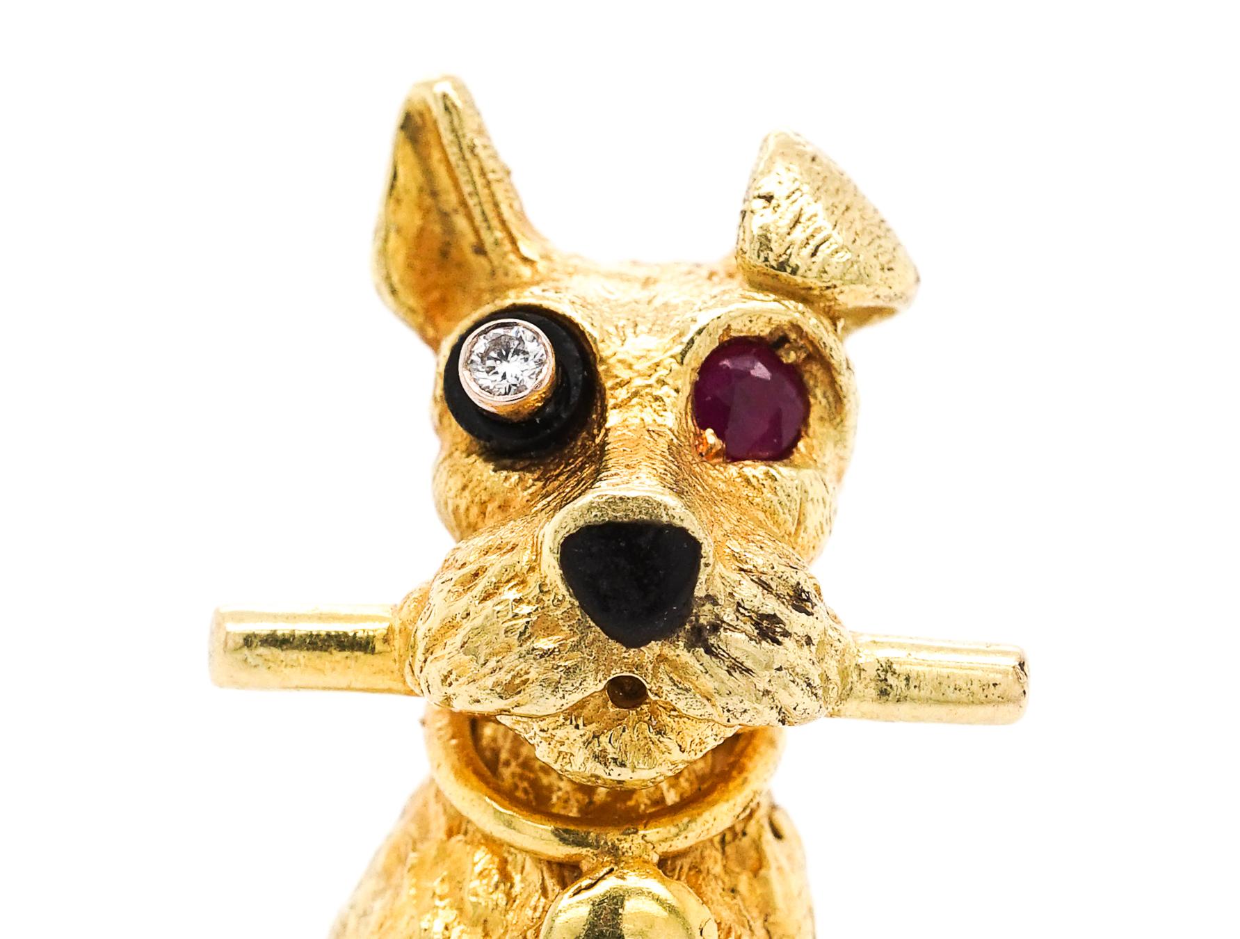 This beautiful 18KT furry terrier clenching a polished stick, with one round diamond eye within a black enamel cylinder, one round ruby eye and black enamel nose, signed Cartier, Paris, no 73, with maker mark. 